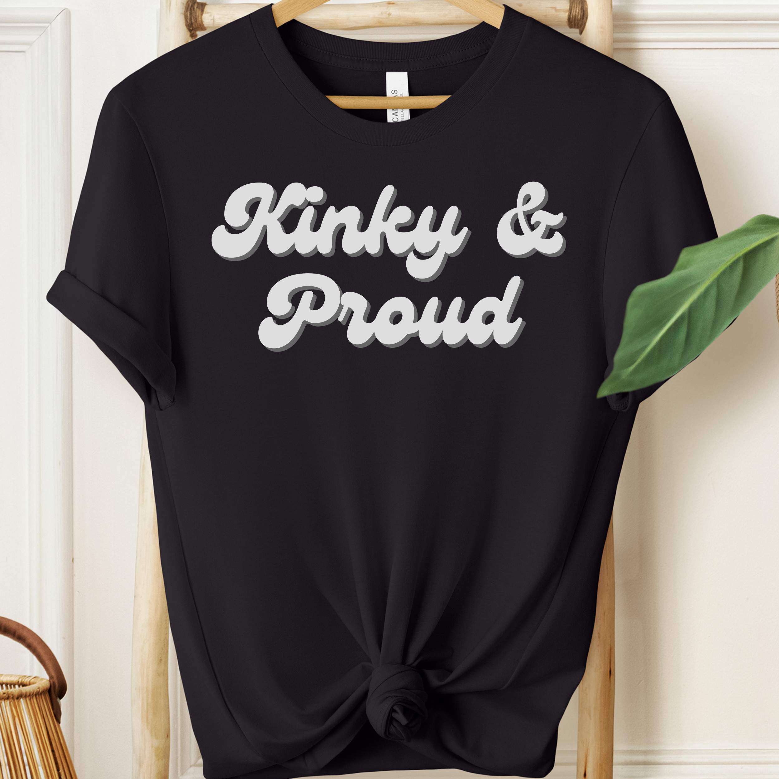 Black t-shirt with 'Kinky & Proud' text on a hanger against a white wall.