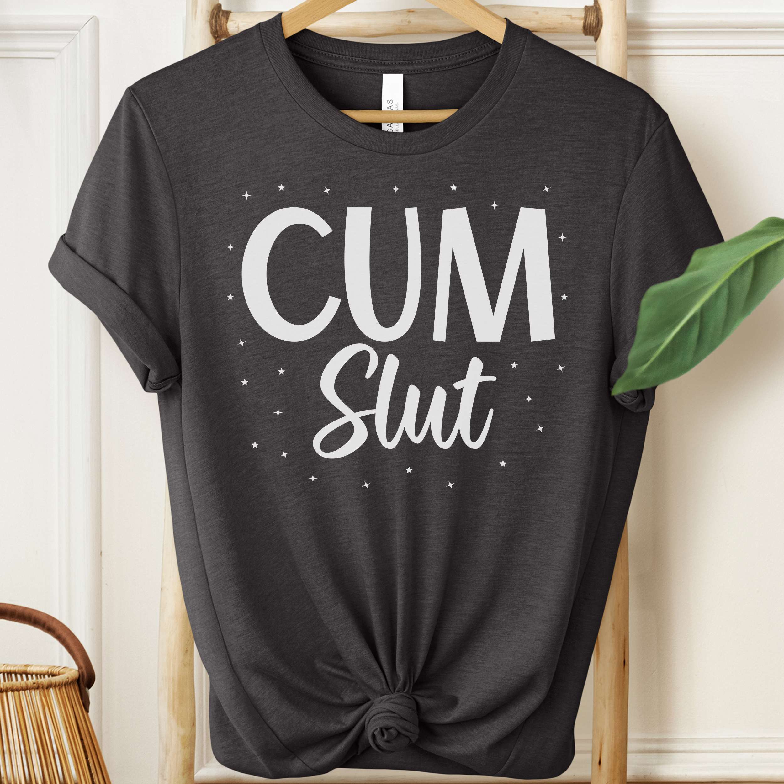 Black t-shirt with 'Cum Slut' text on a hanger against a white wall.