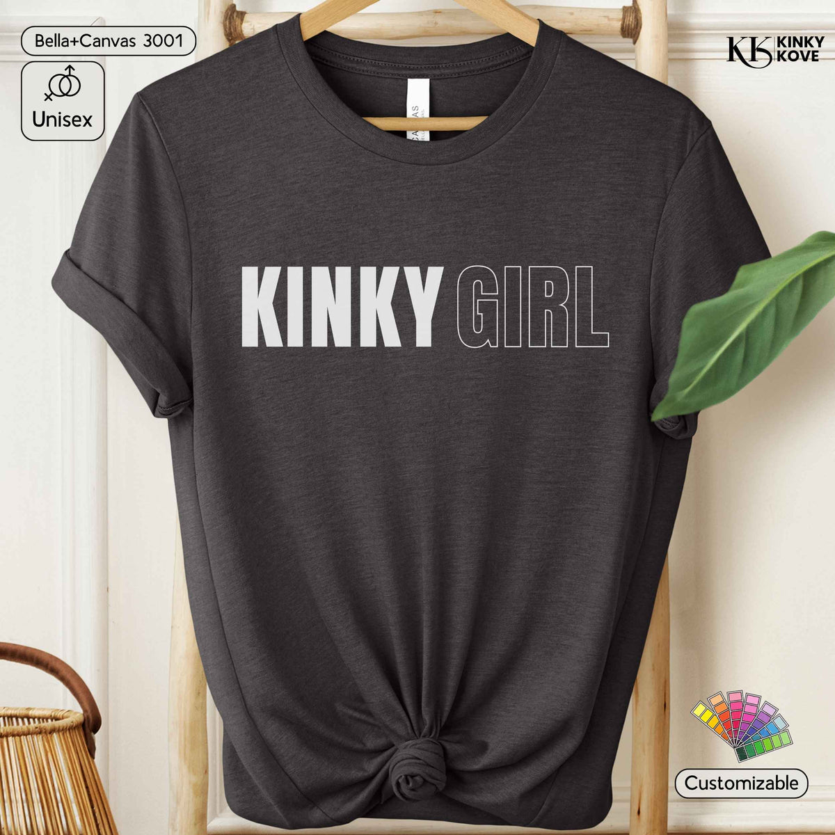 A dark charcoal gray t-shirt with rolled sleeves and a knotted hem, featuring the text 'KINKY GIRL' printed across the chest in white. The shirt is displayed on a wooden hanger against a light cream background with decorative props including a green leaf, a wicker basket, and branding overlays.