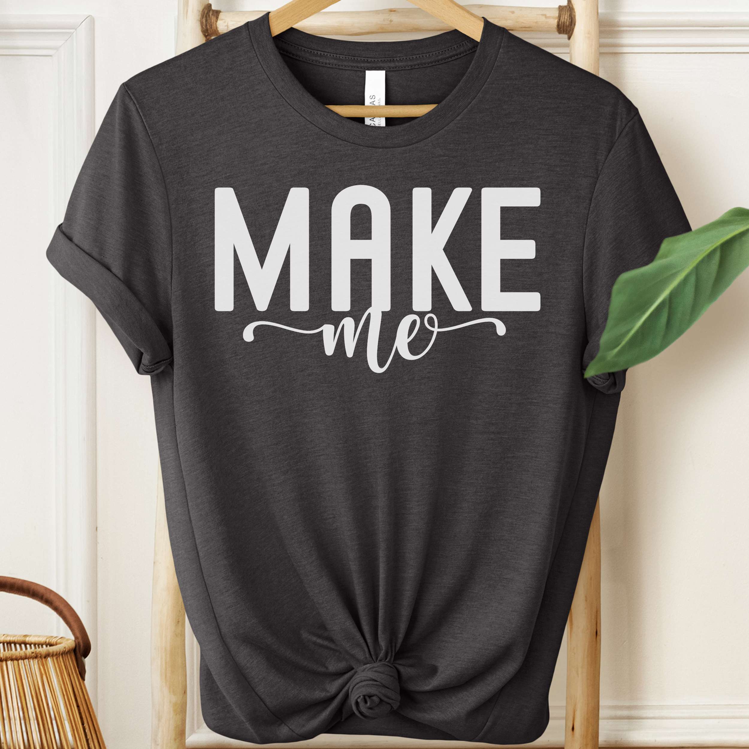 Black t-shirt with 'MAKE me' text on a hanger against a white wall.