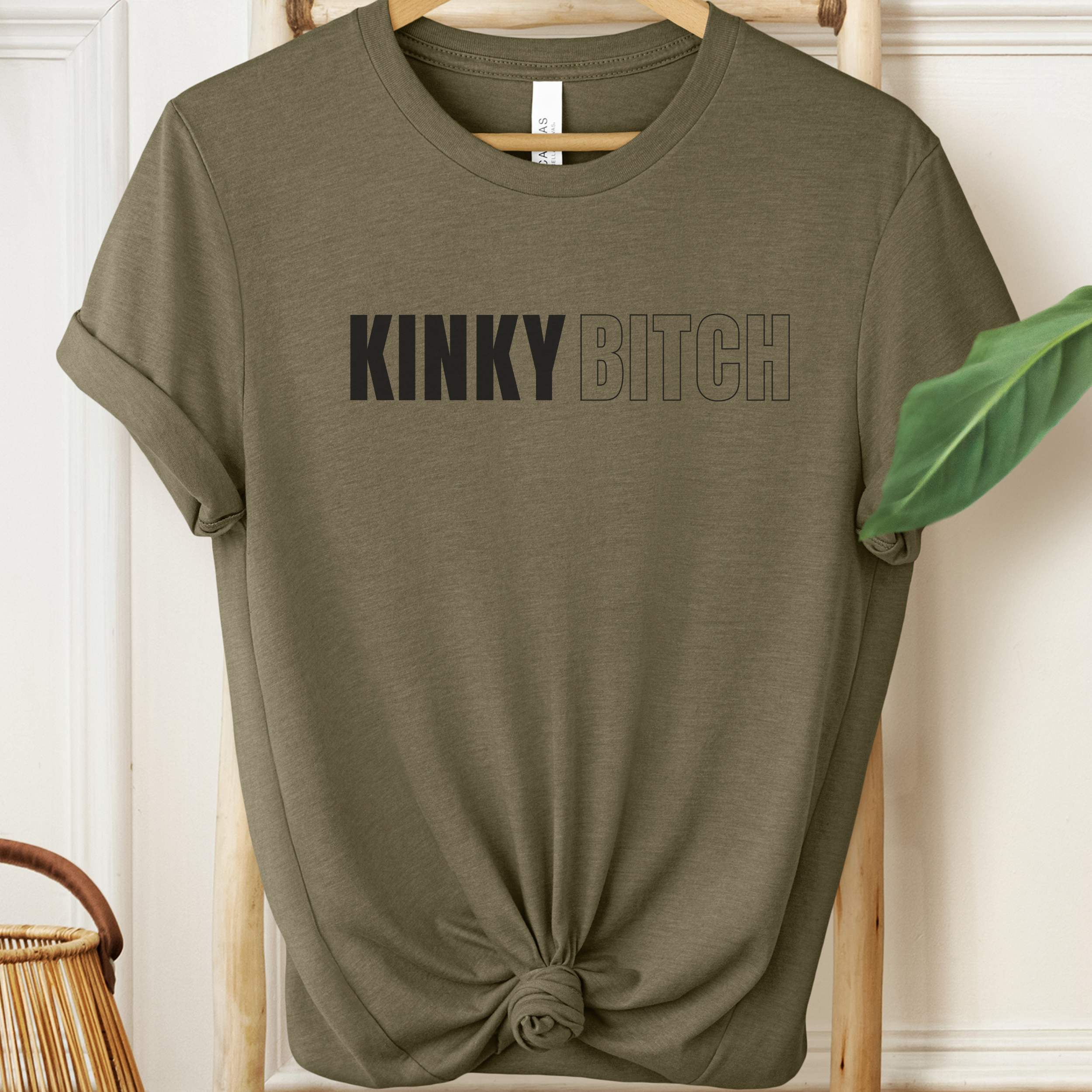 Olive green t-shirt with 'KINKY BITCH' text on a wooden hanger.