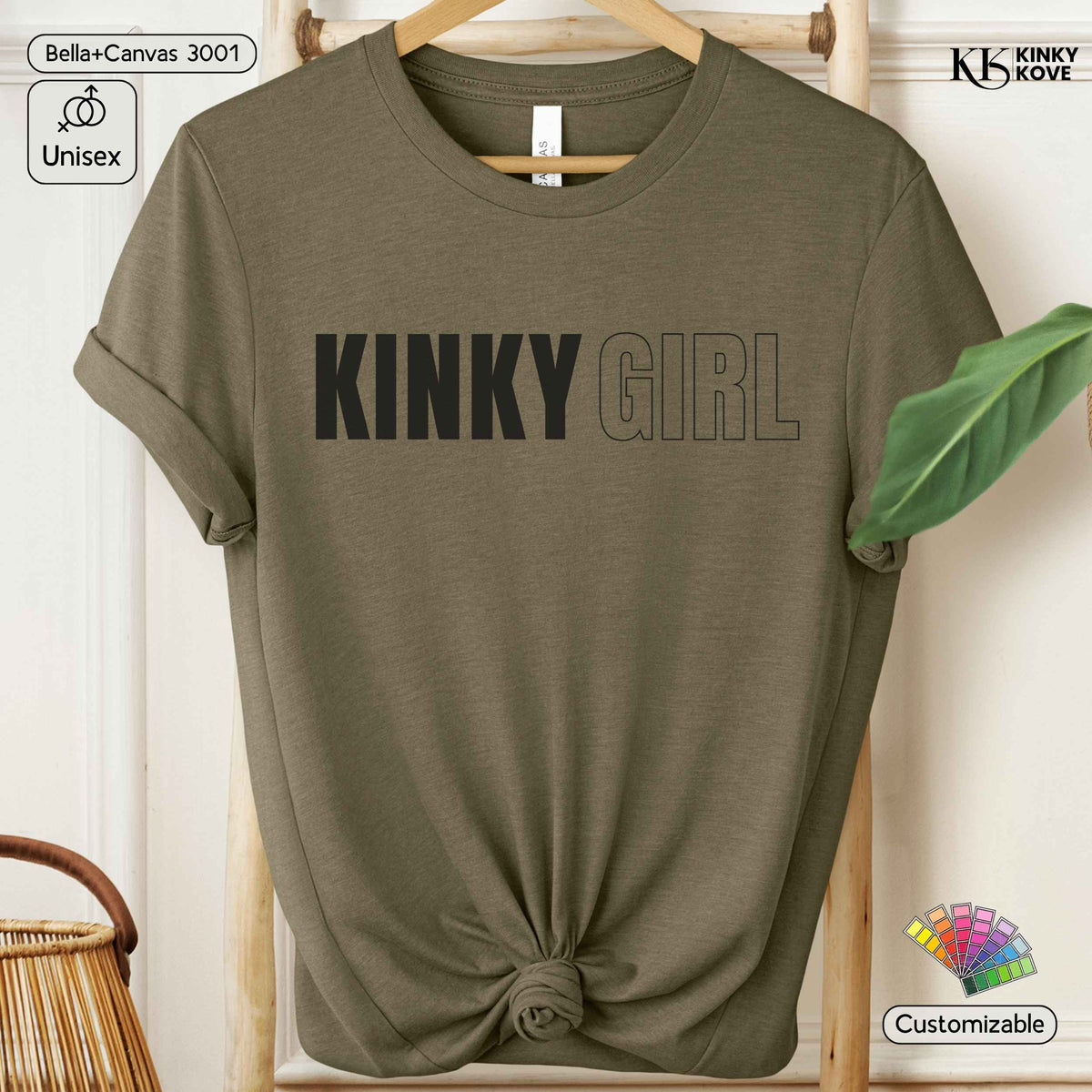 An olive green short-sleeve t-shirt with 'KINKY GIRL' printed across the chest, styled on a wooden hanger with a knotted hem, set against a light wood background with decorative props.
