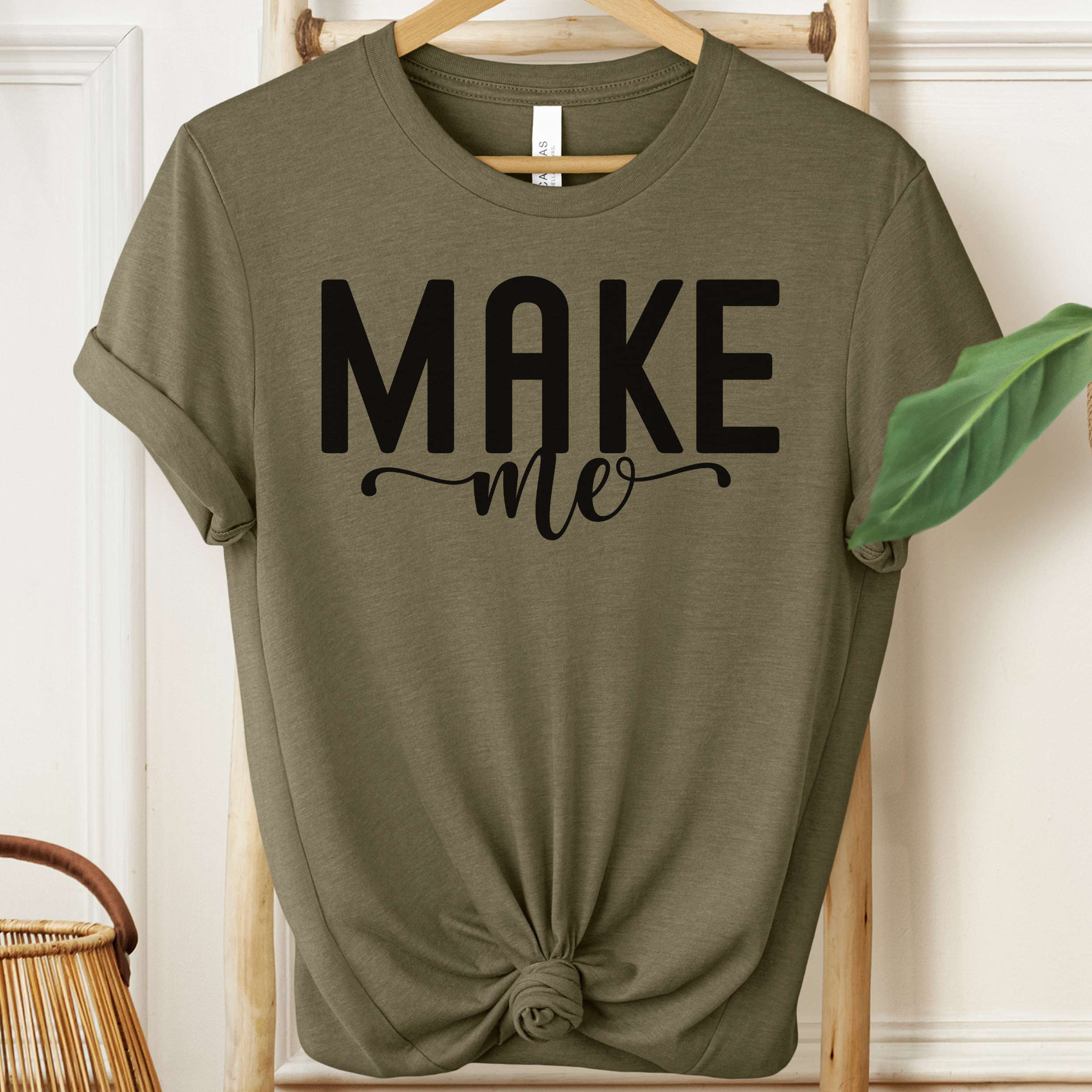 Green t-shirt with 'MAKE me' text on a wooden hanger against a white wall.