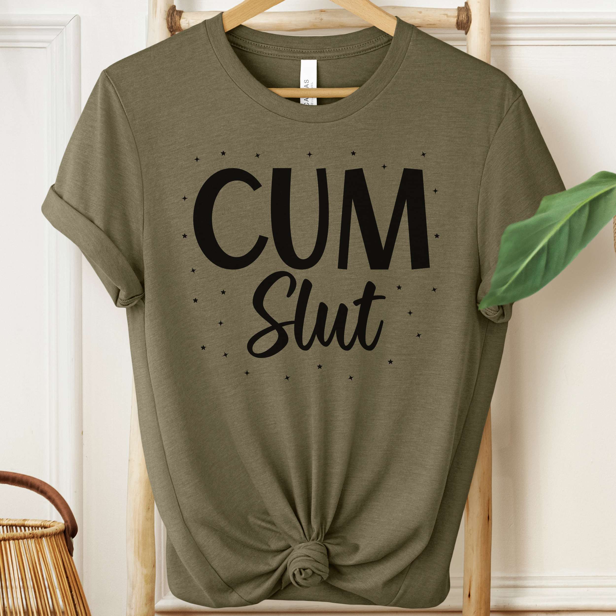 Green t-shirt with 'Cum Slut' text on a hanger against a white wall.