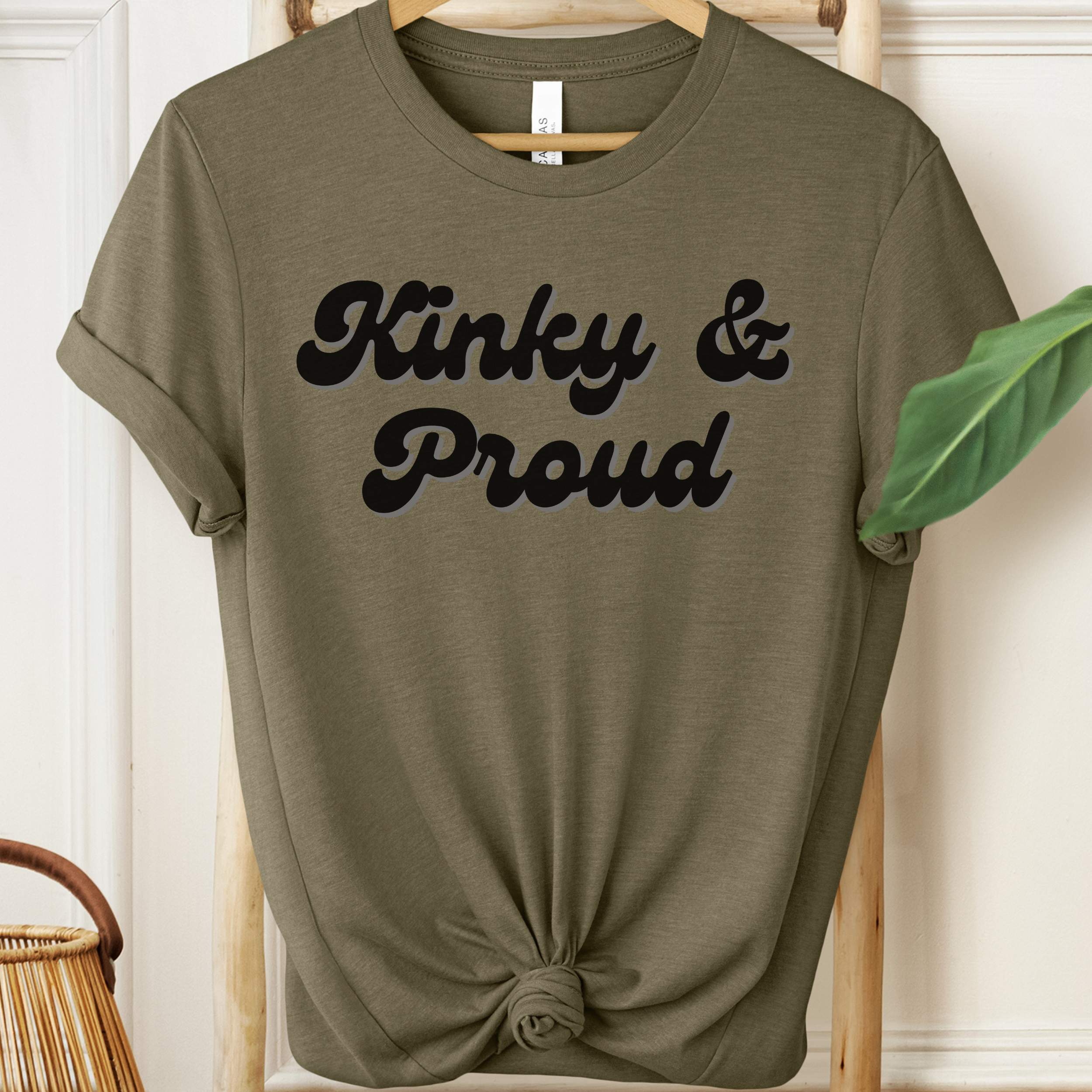 Olive green t-shirt with 'Kinky & Proud' text on a hanger.
