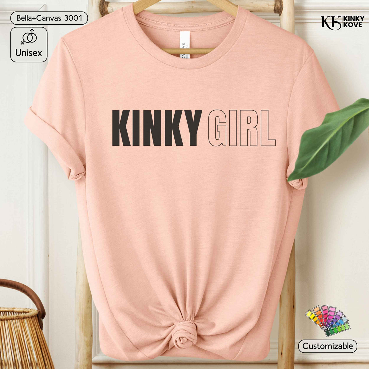 A light pink short-sleeve t-shirt with 'KINKY GIRL' printed across the chest, styled on a wooden hanger with a knot at the bottom hem, set against a neutral indoor background with decorative props.