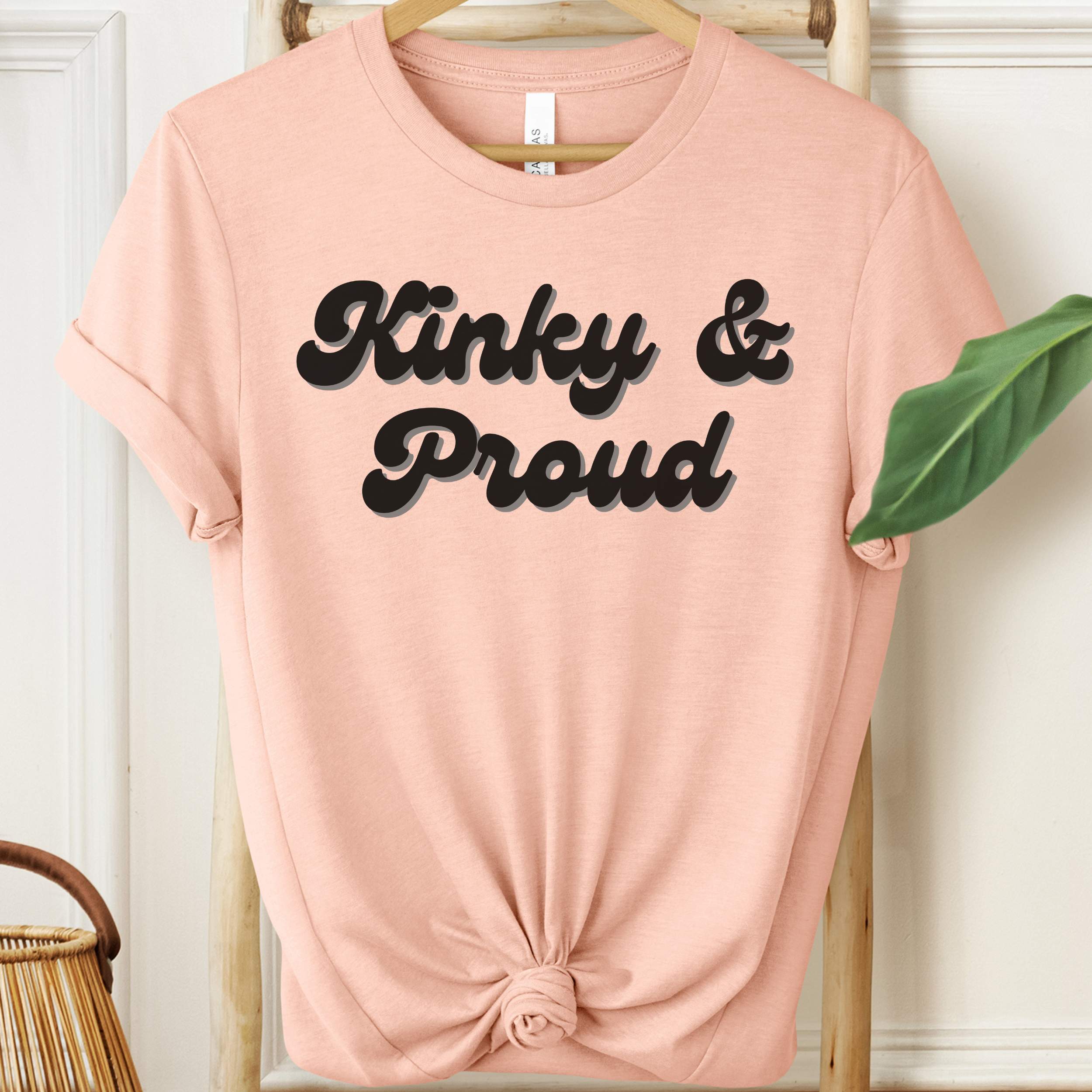 Pink t-shirt with 'Kinky & Proud' text on a hanger against a white wall.