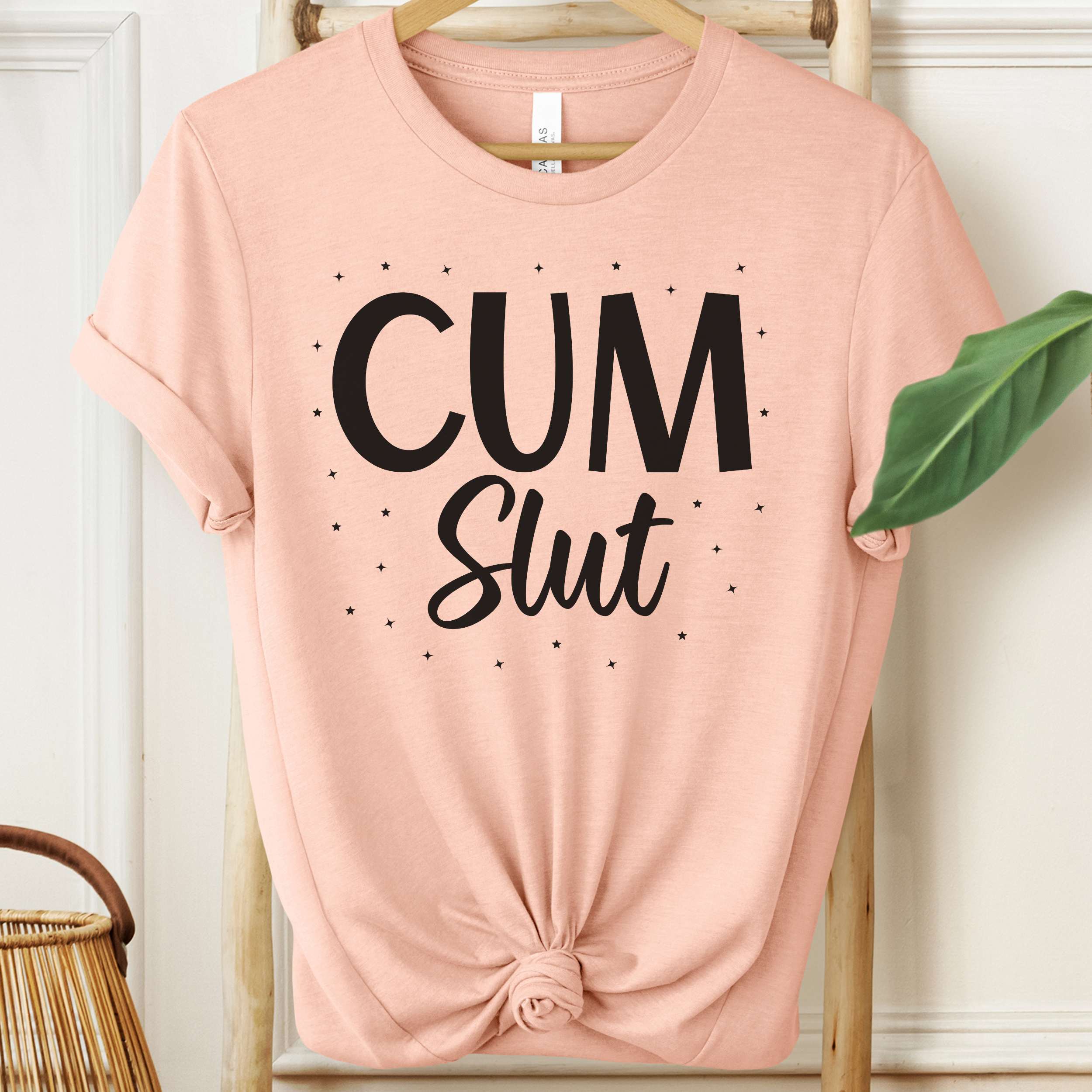 Pink t-shirt with 'Cum Slut' text on a hanger against a white wall.