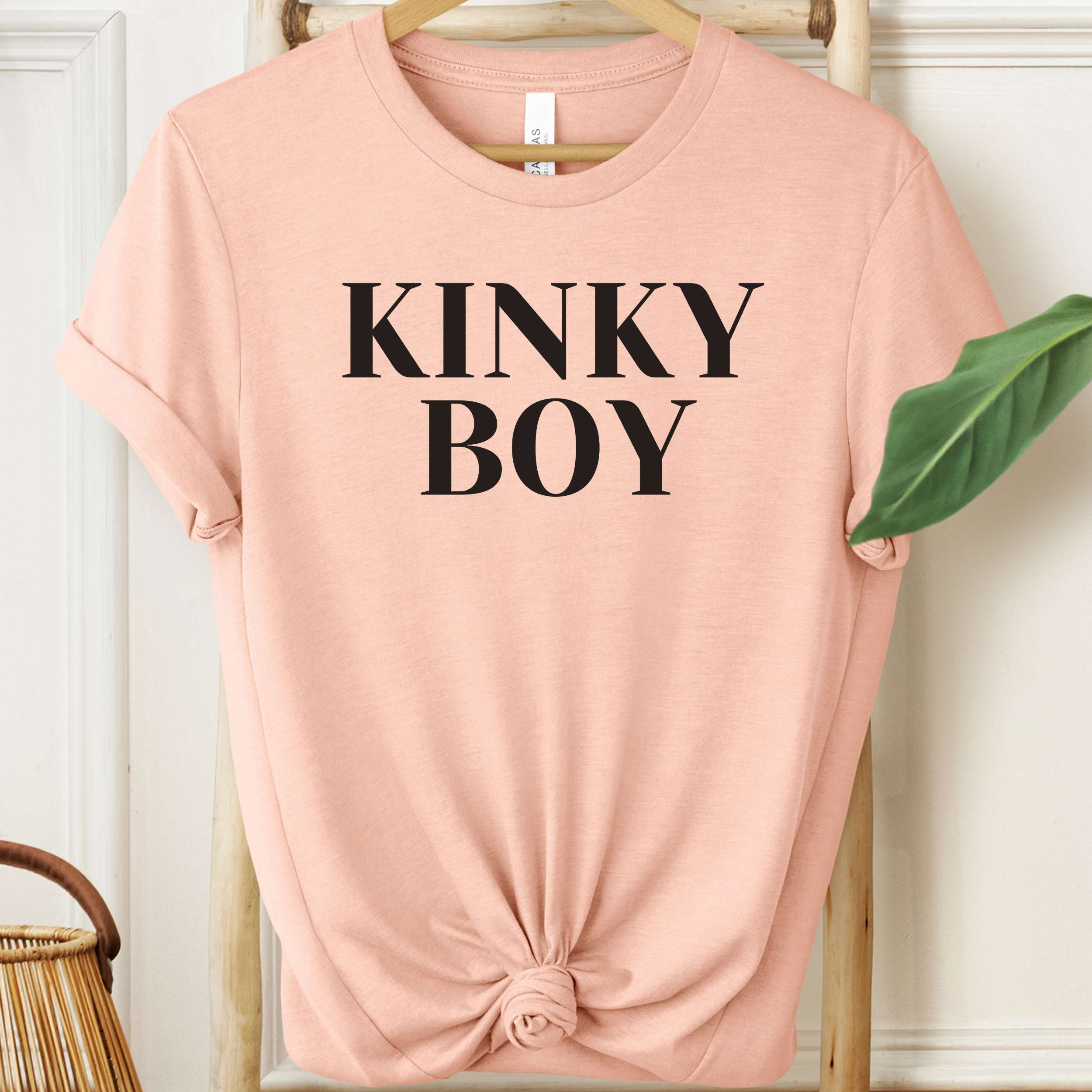 Pink t-shirt with 'KINKY BOY' text on a hanger against a white wall.