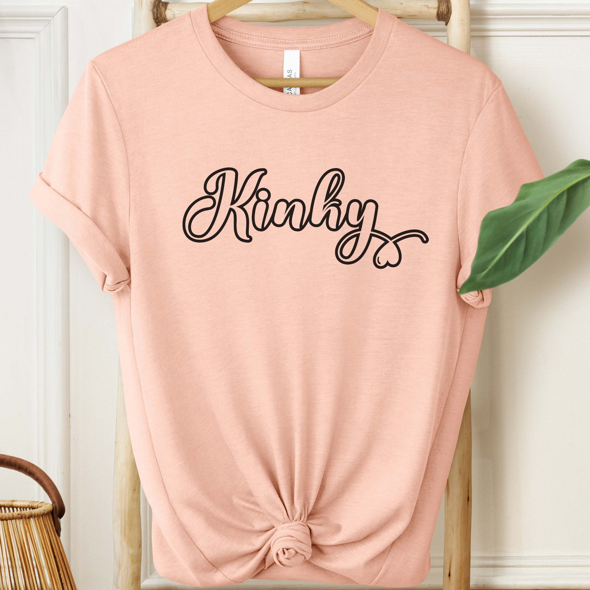 Pink t-shirt with 'Kinky' text on a hanger against a white wall.