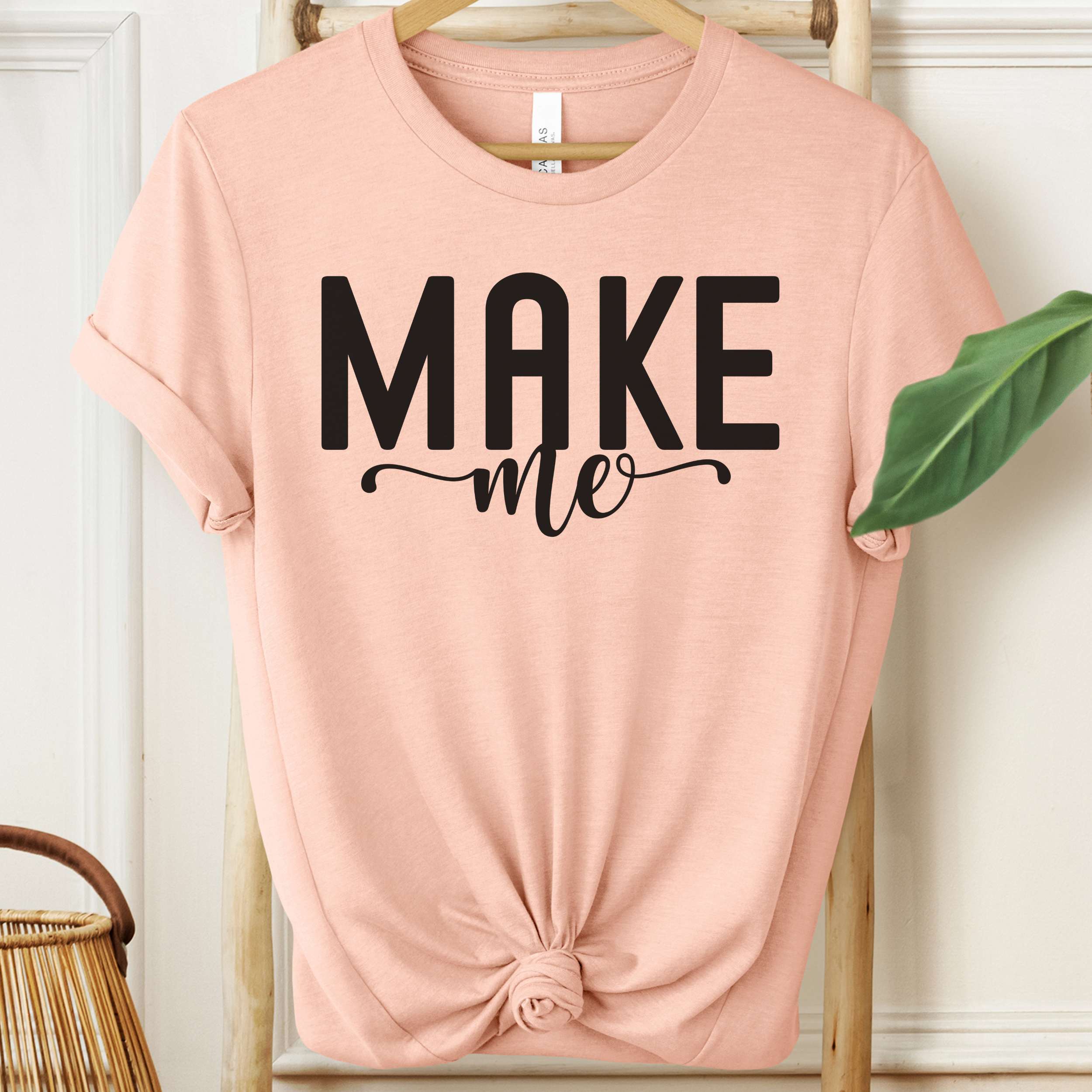 Pink t-shirt with 'MAKE me' text on a hanger against a white wall.