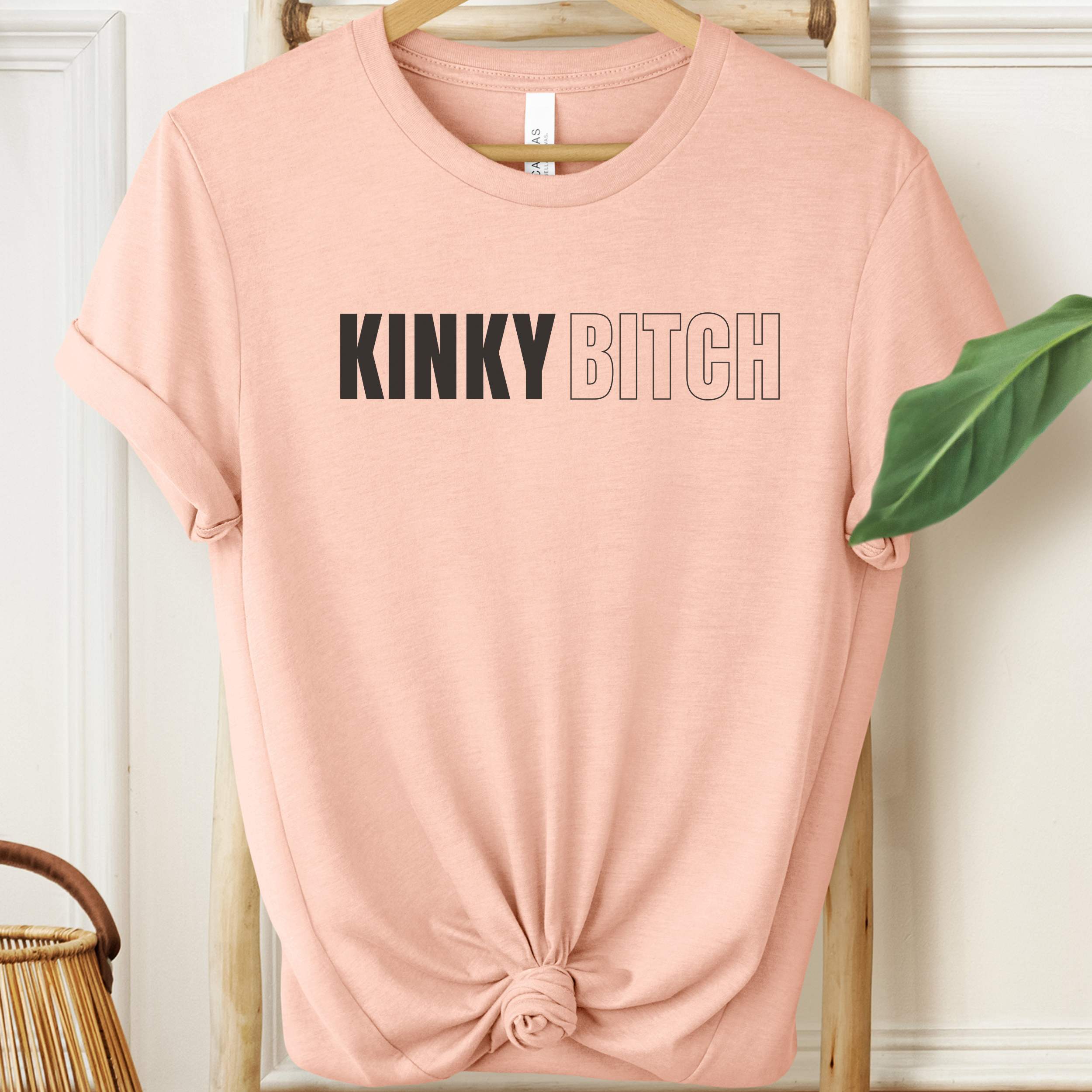 Pink t-shirt with 'KINKY BITCH' text on a hanger against a white wall.