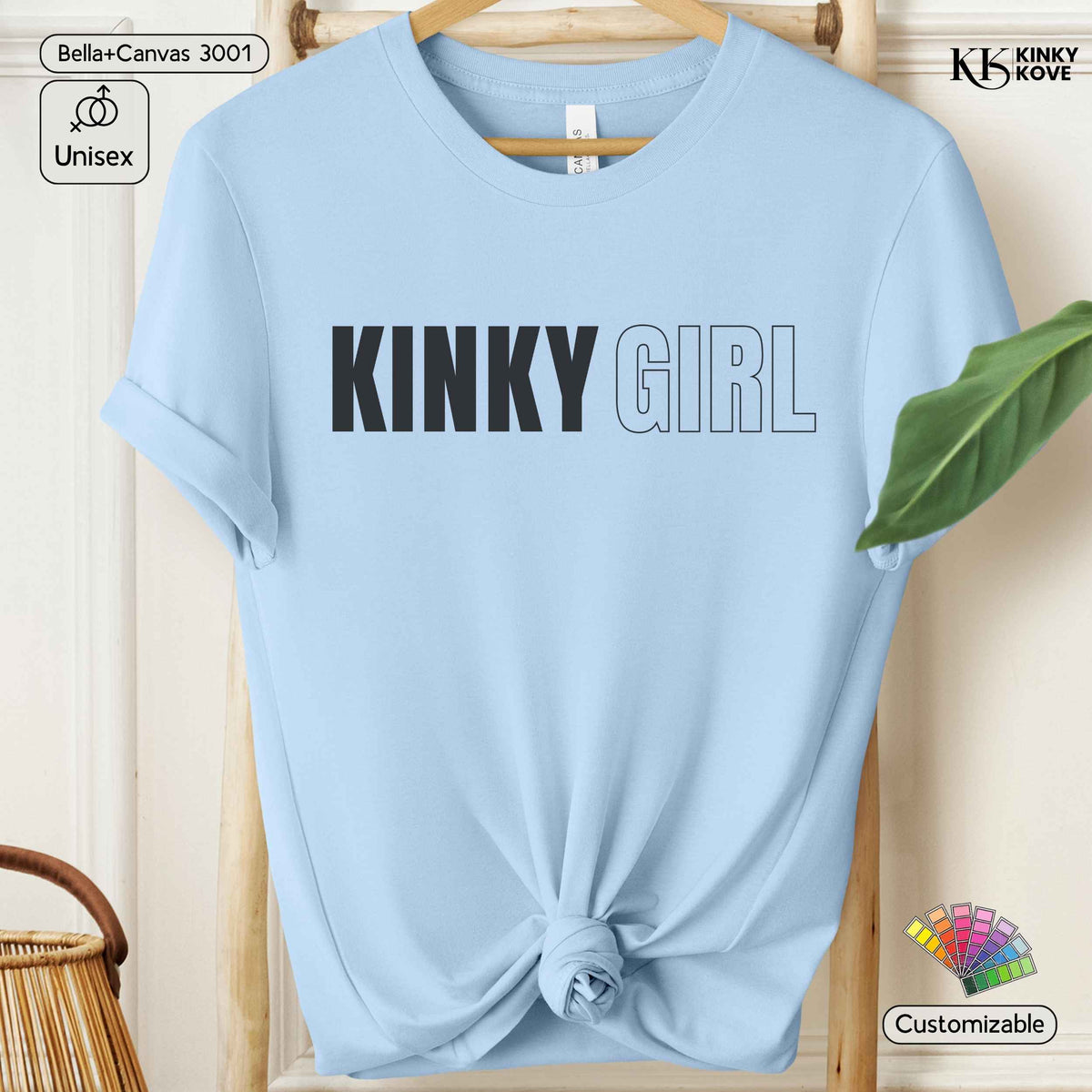 A light blue short-sleeve t-shirt with 'KINKY GIRL' printed across the chest, styled on a wooden hanger with a knot at the bottom hem. The background shows a light wood wall and a green leaf.