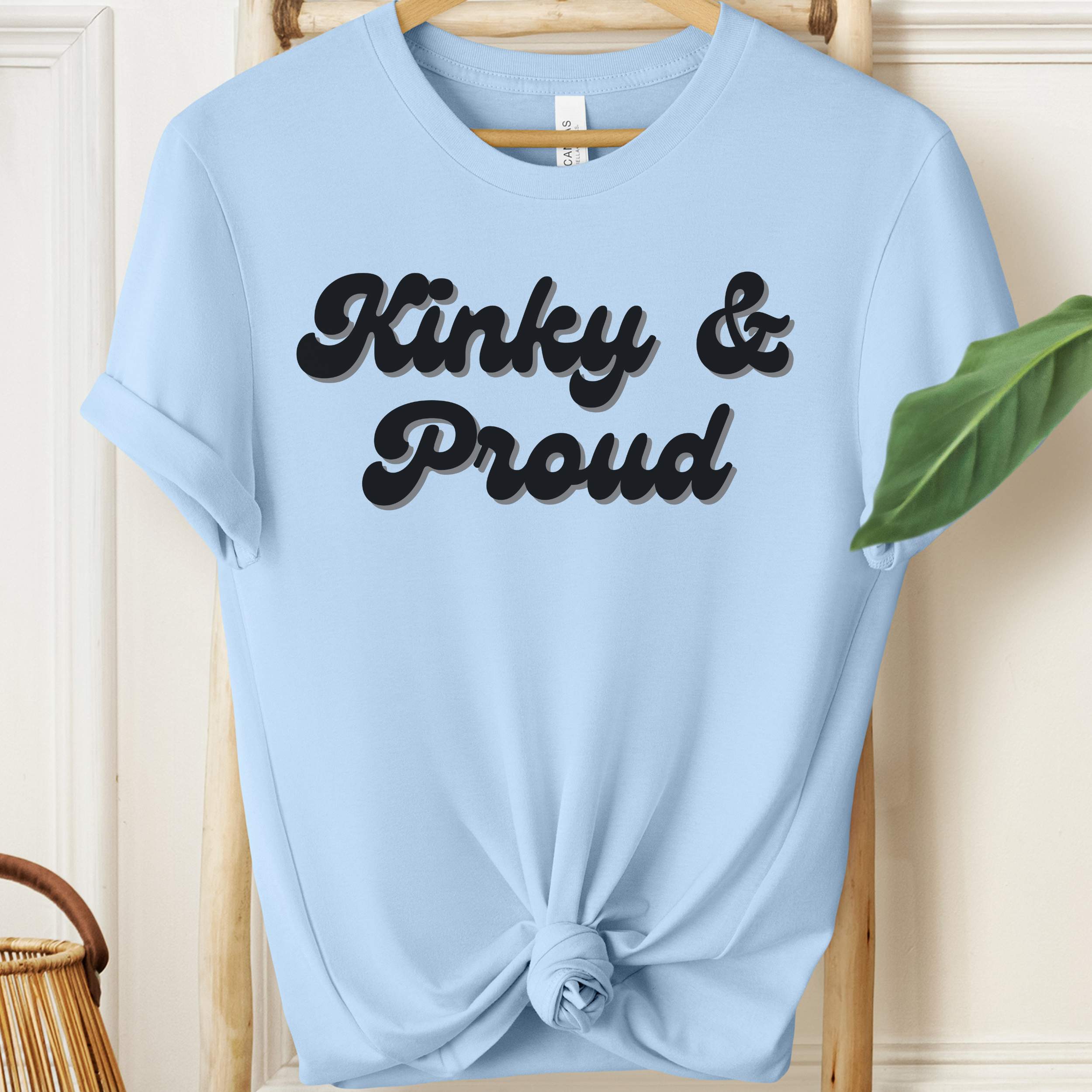 Light blue t-shirt with 'Kinky & Proud' text on a wooden hanger.
