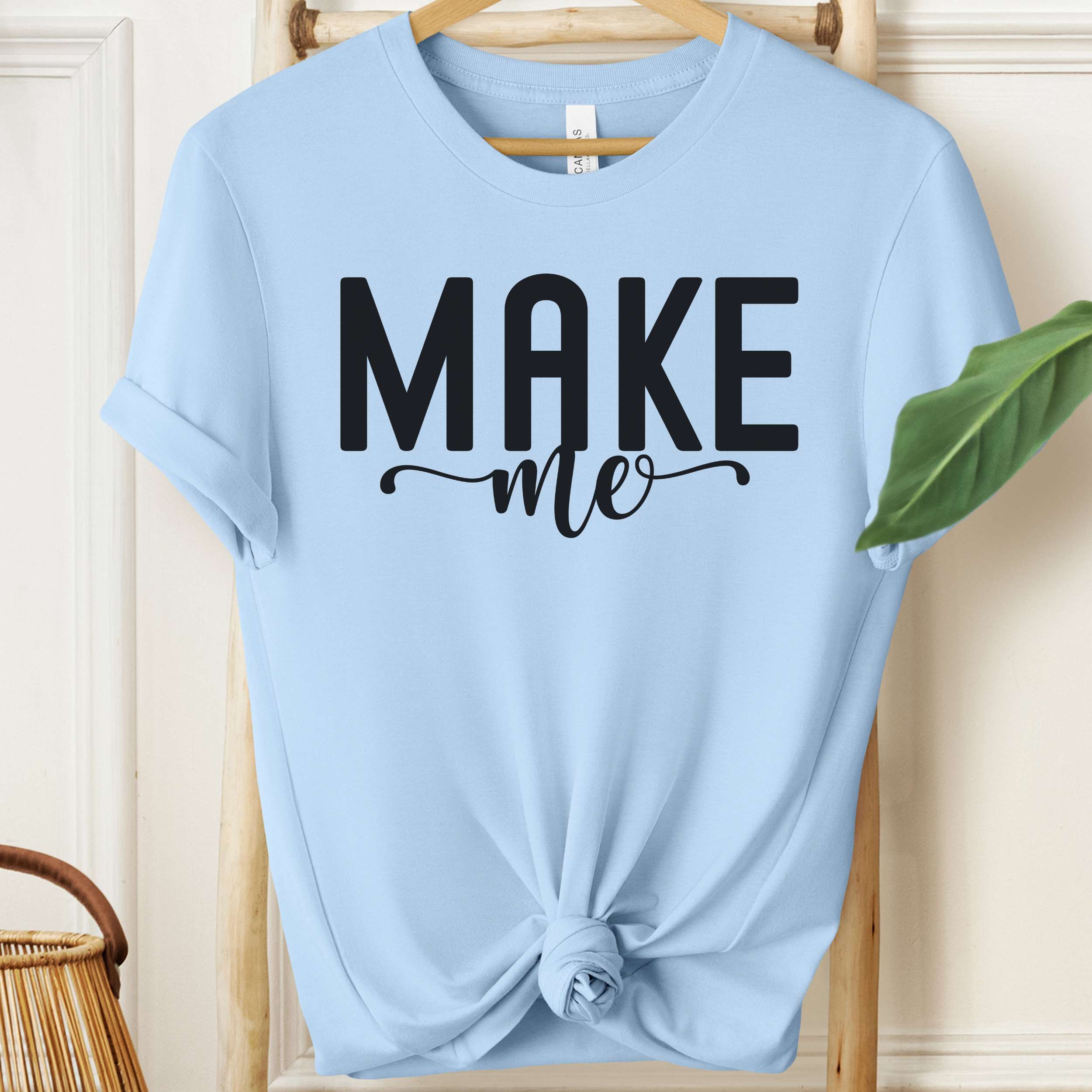 Light blue t-shirt with 'MAKE me' text on a wooden hanger.