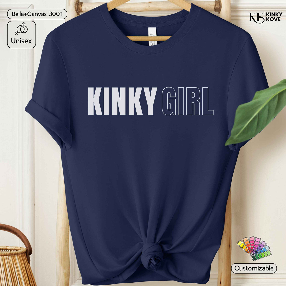 A navy blue short-sleeve t-shirt with 'KINKY GIRL' printed across the chest in white and outlined text, styled on a wooden hanger with a knotted hem. The image includes decorative props like a green leaf and a wicker basket, along with several digital overlay badges.
