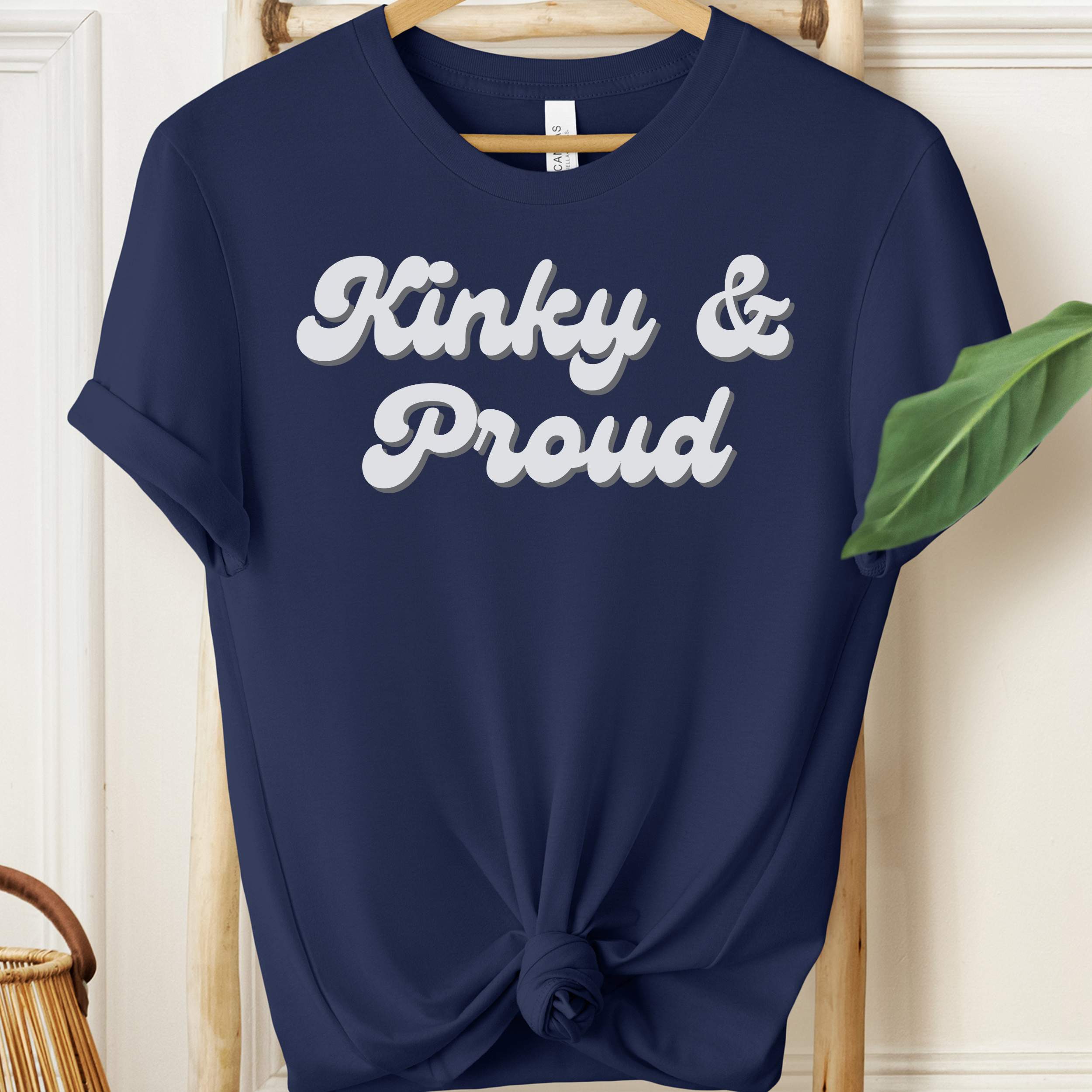 Navy blue t-shirt with 'Kinky & Proud' text on a wooden hanger.