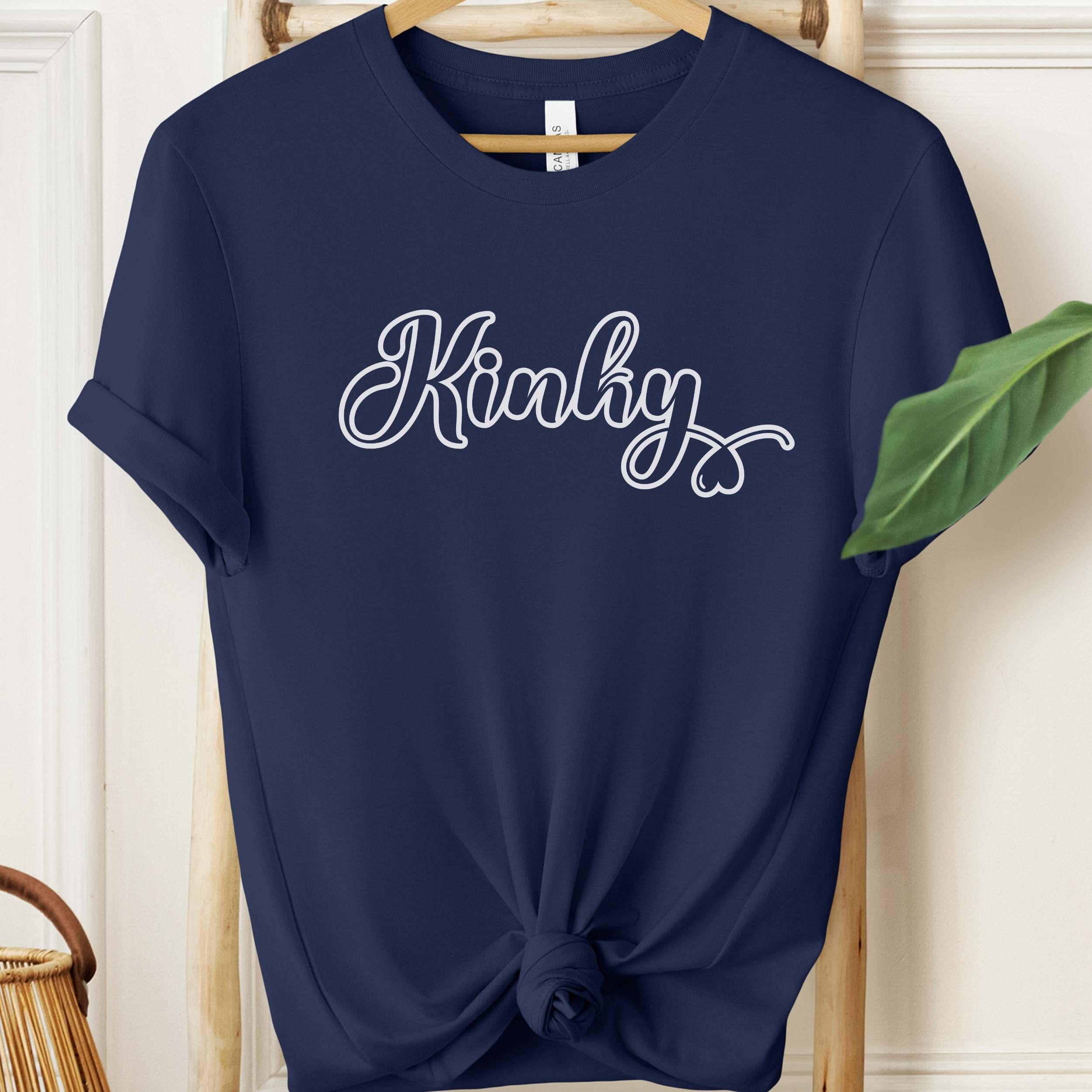 Navy blue t-shirt with 'Kinky' text on a wooden hanger against a white wall.