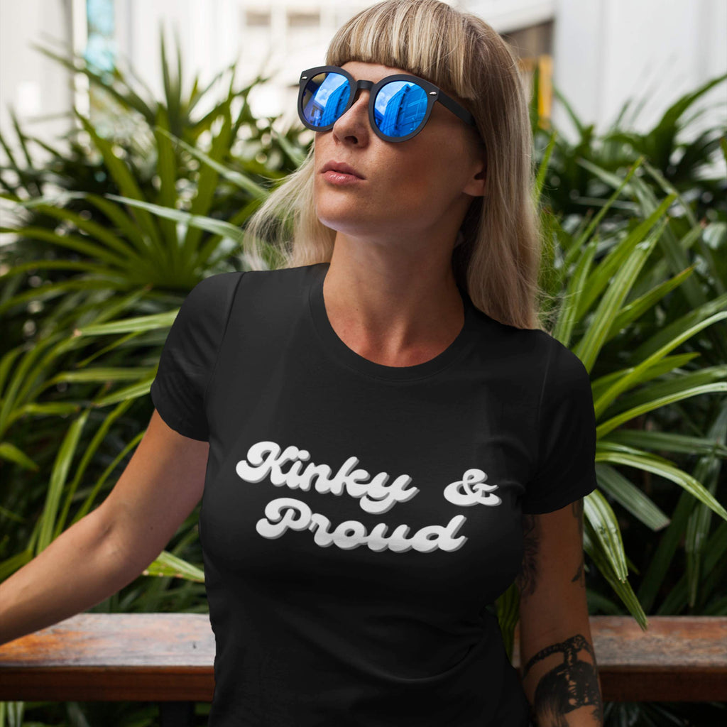Woman wearing a black t-shirt with 'Kinky & Proud' text, standing outdoors with greenery in the background.