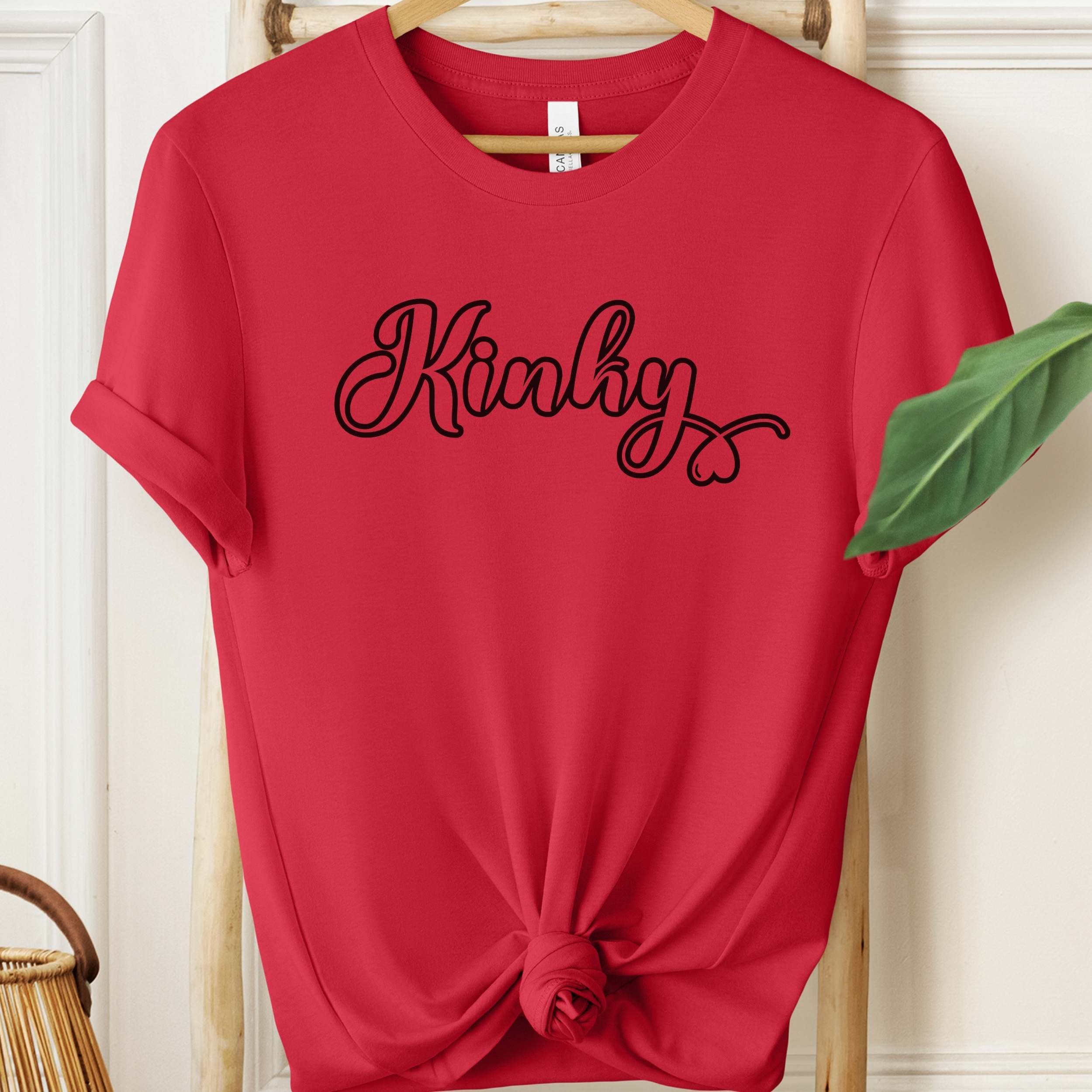 Red t-shirt with 'Kinky' text on a hanger against a white wall.