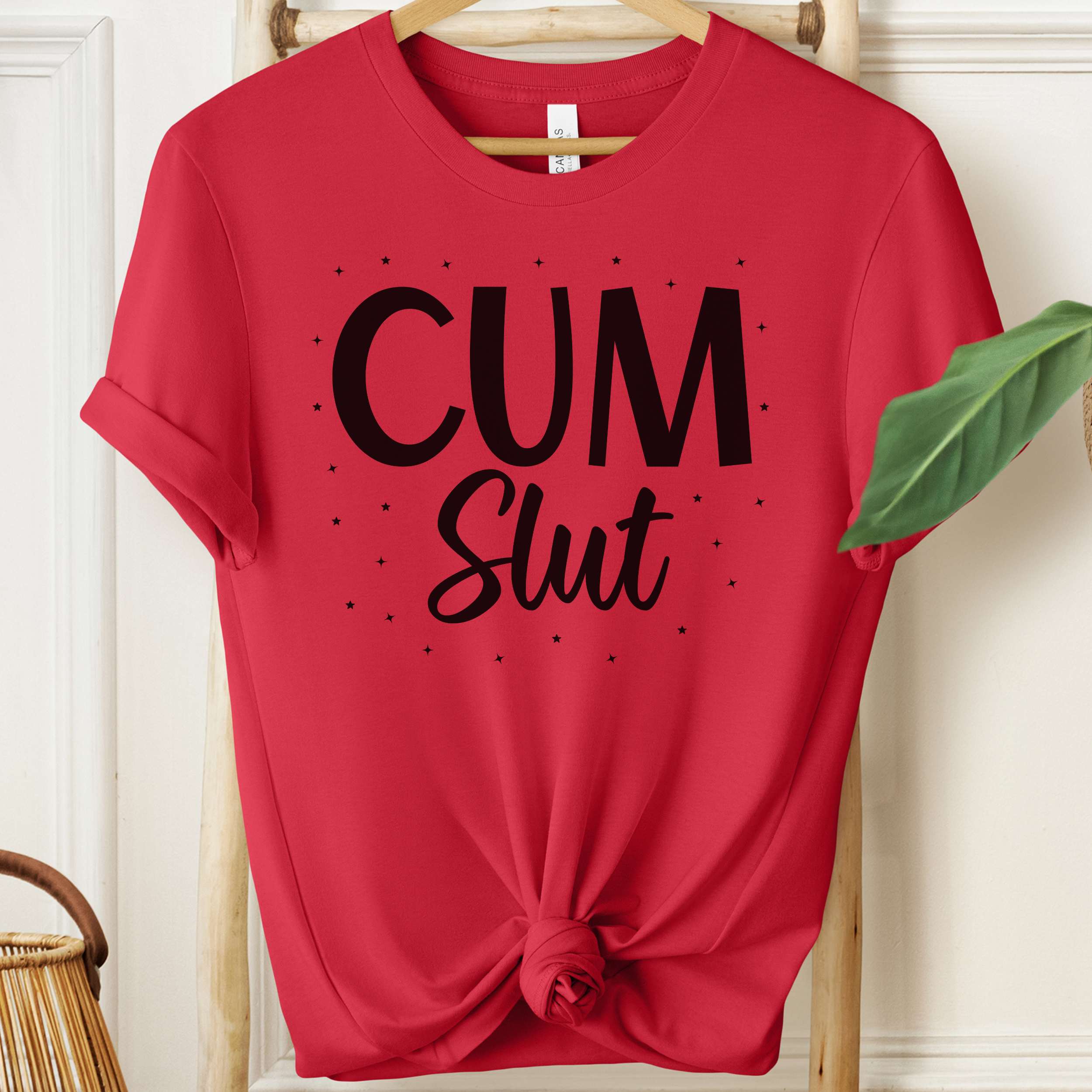 Red t-shirt with 'Cum Slut' text on a hanger against a white wall.