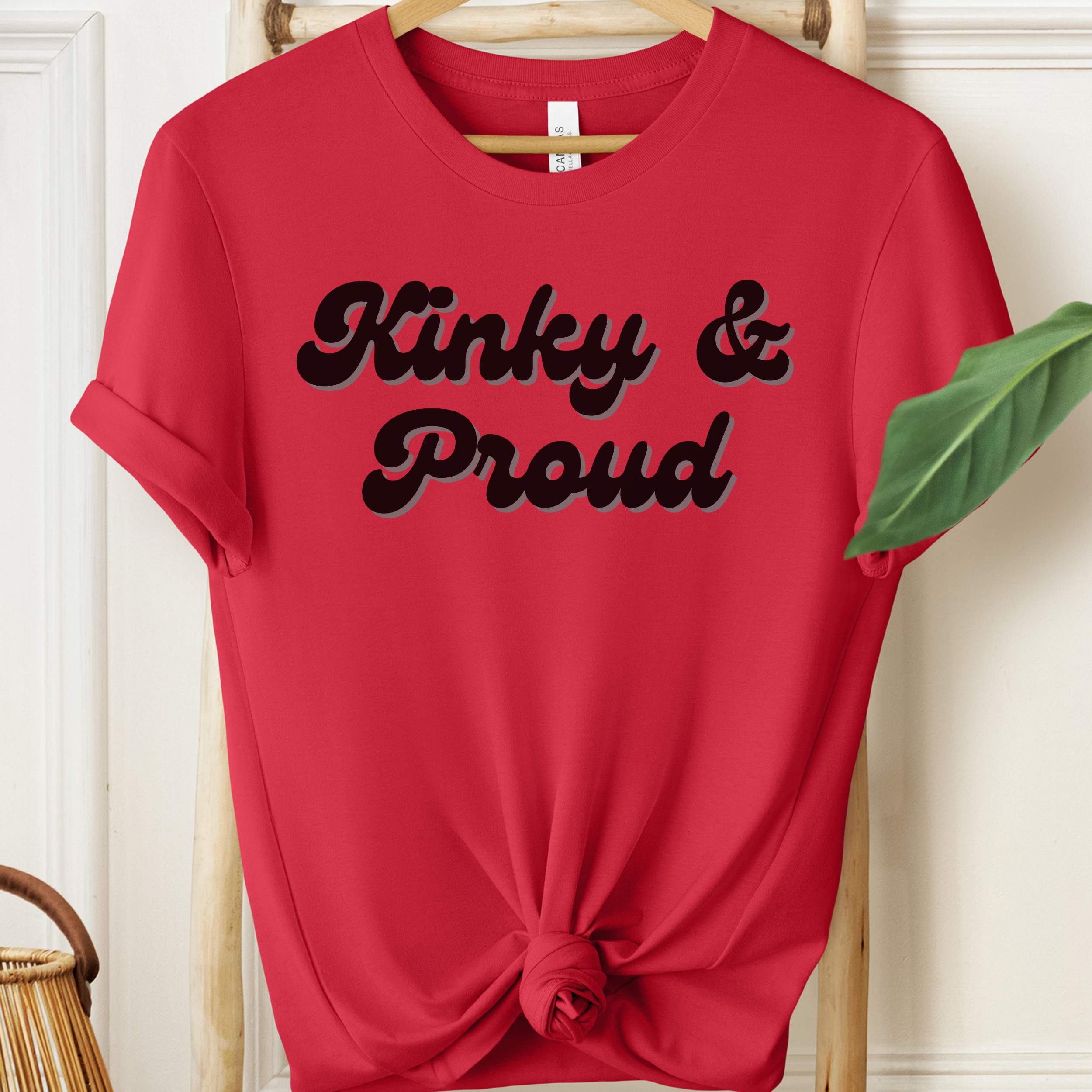 Red t-shirt with 'Kinky & Proud' text on a hanger against a white wall.