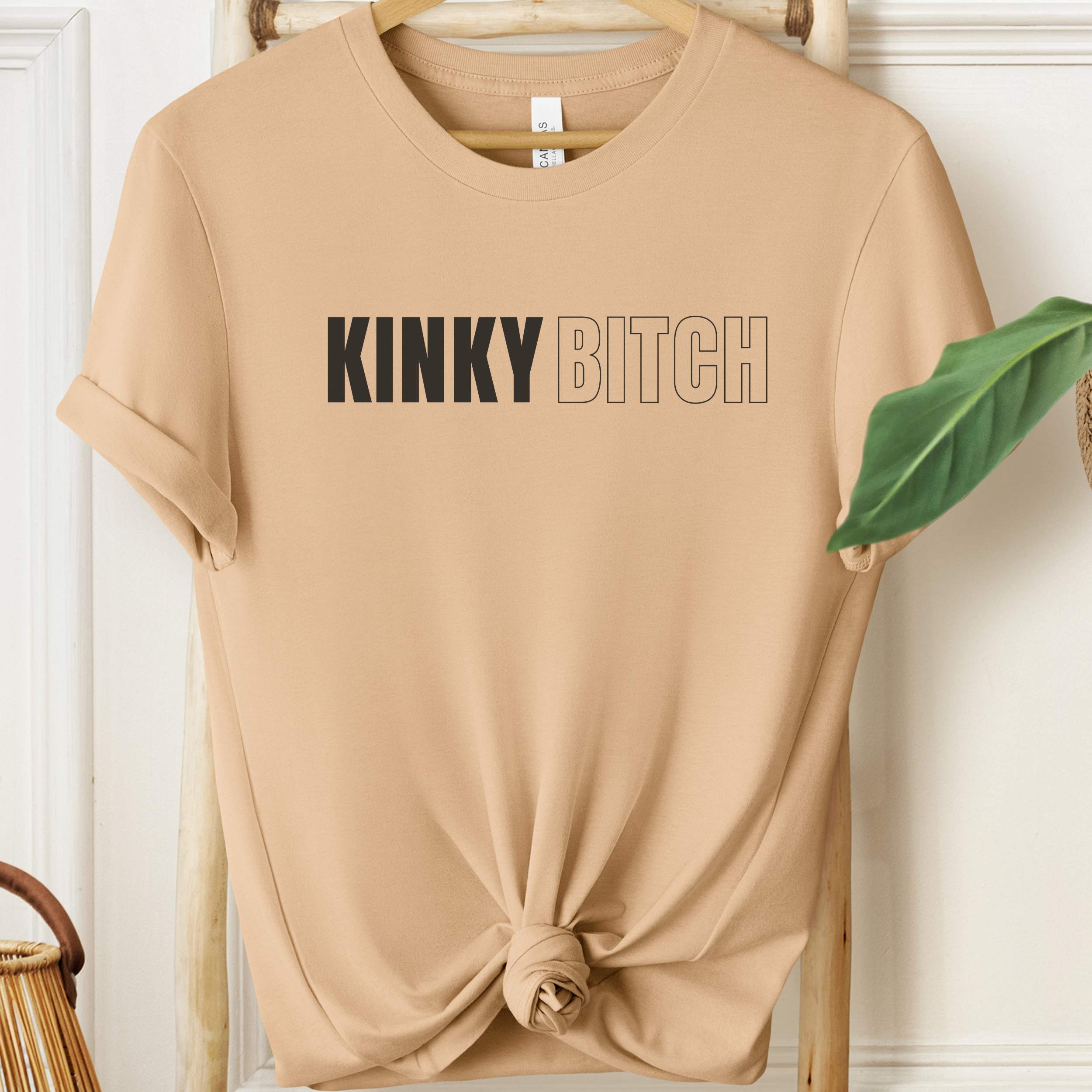 Beige t-shirt with 'KINKY BITCH' text on a wooden hanger.