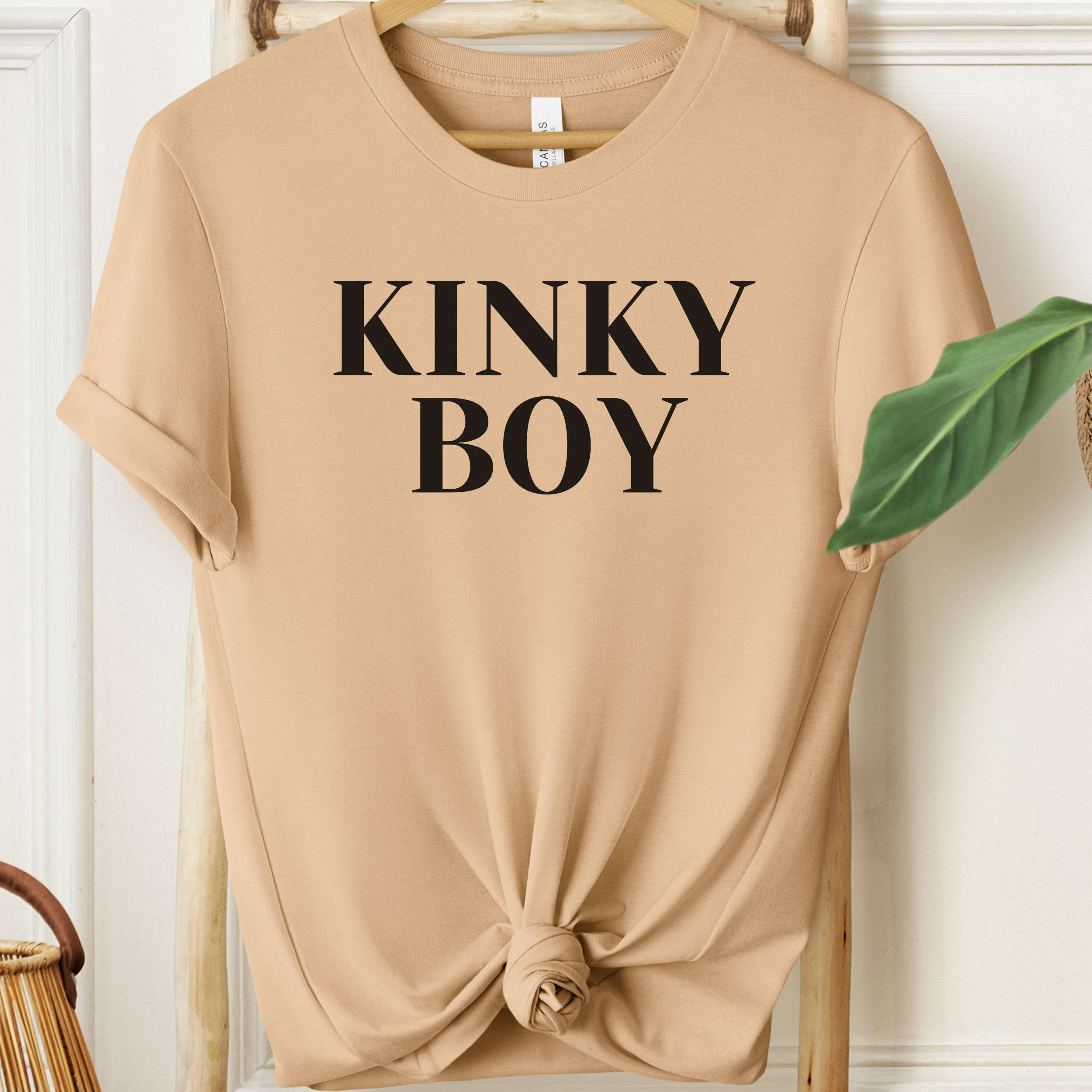 Beige t-shirt with 'KINKY BOY' text on a wooden hanger against a white wall.
