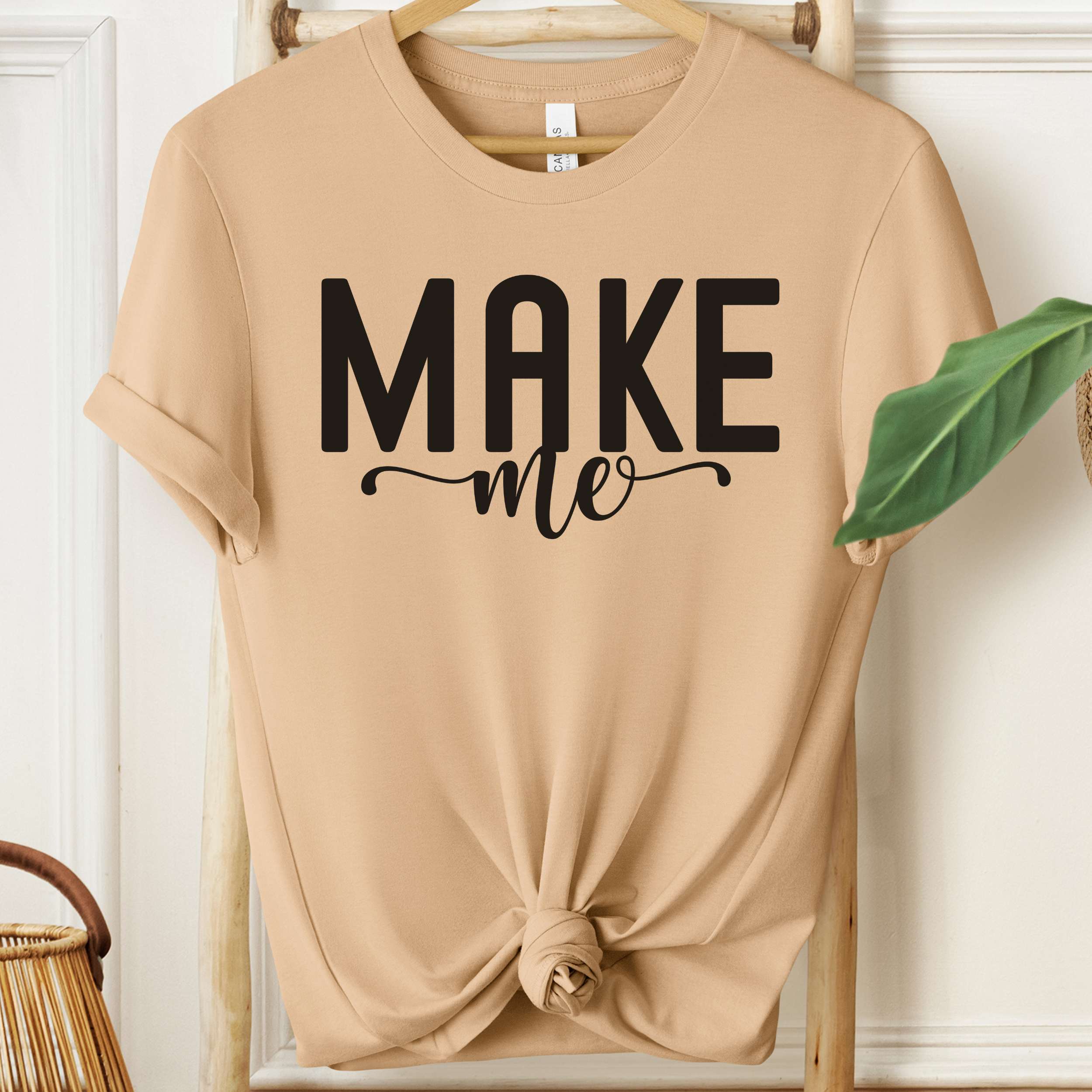 Beige t-shirt with 'MAKE me' text on a hanger against a white wall.