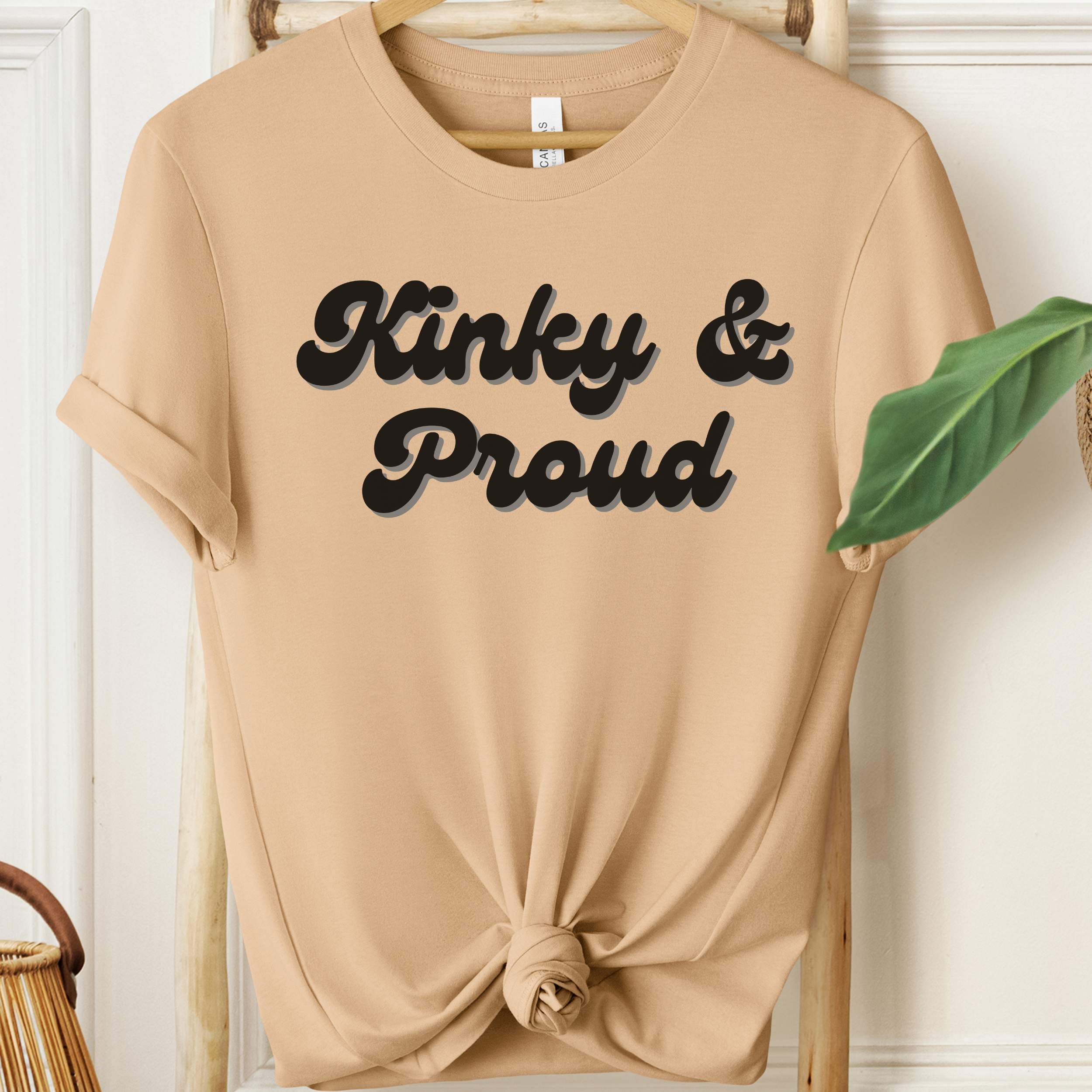 Beige t-shirt with 'Kinky & Proud' text on a hanger against a white wall.