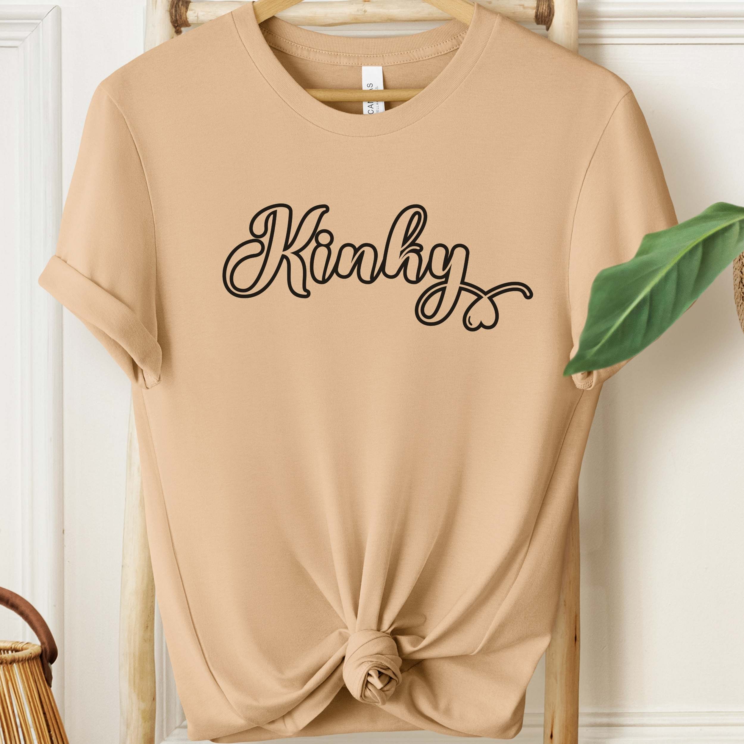 Beige t-shirt with 'Kinky' text on a wooden hanger against a white wall.