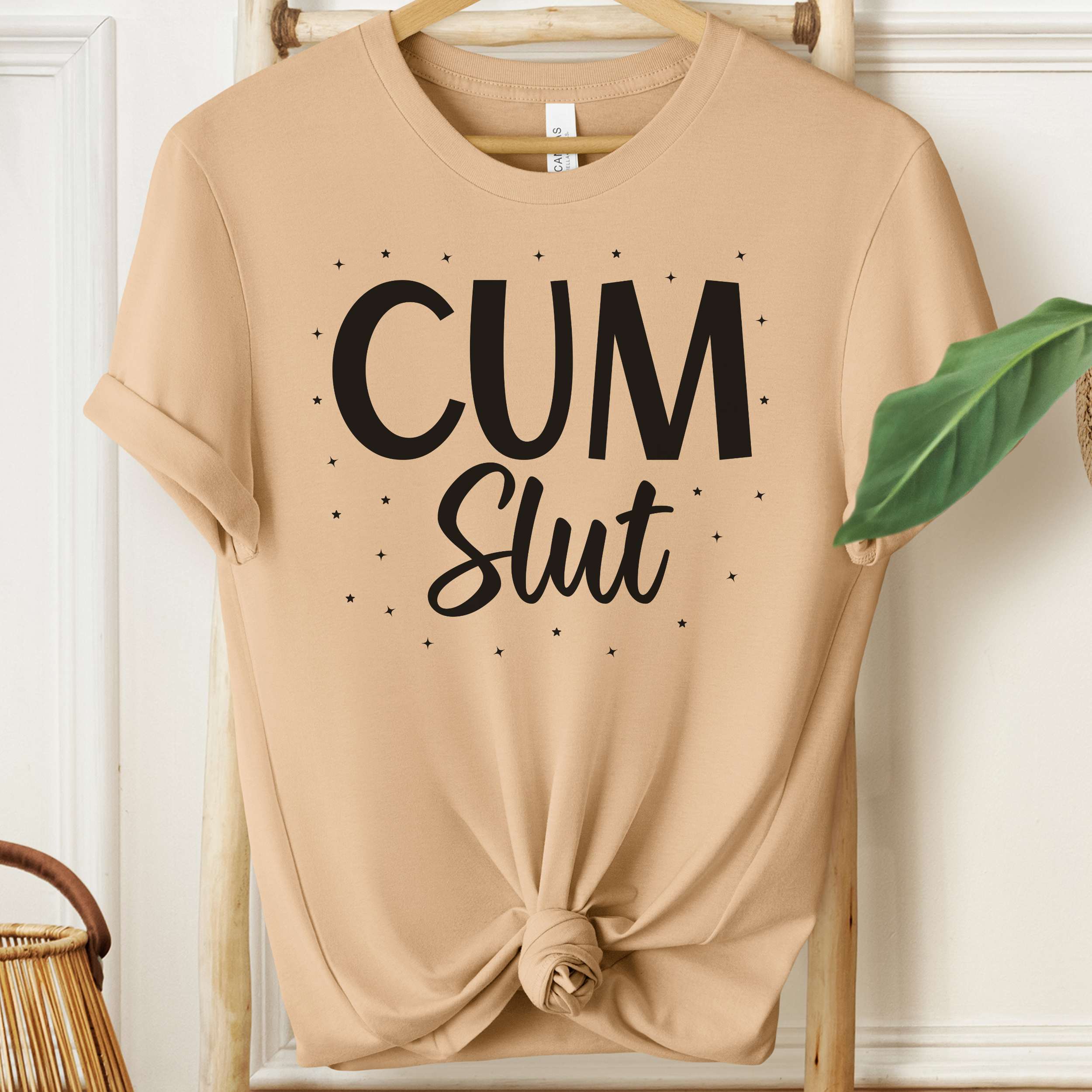 Beige t-shirt with 'Cum Slut' text on a hanger against a white wall.