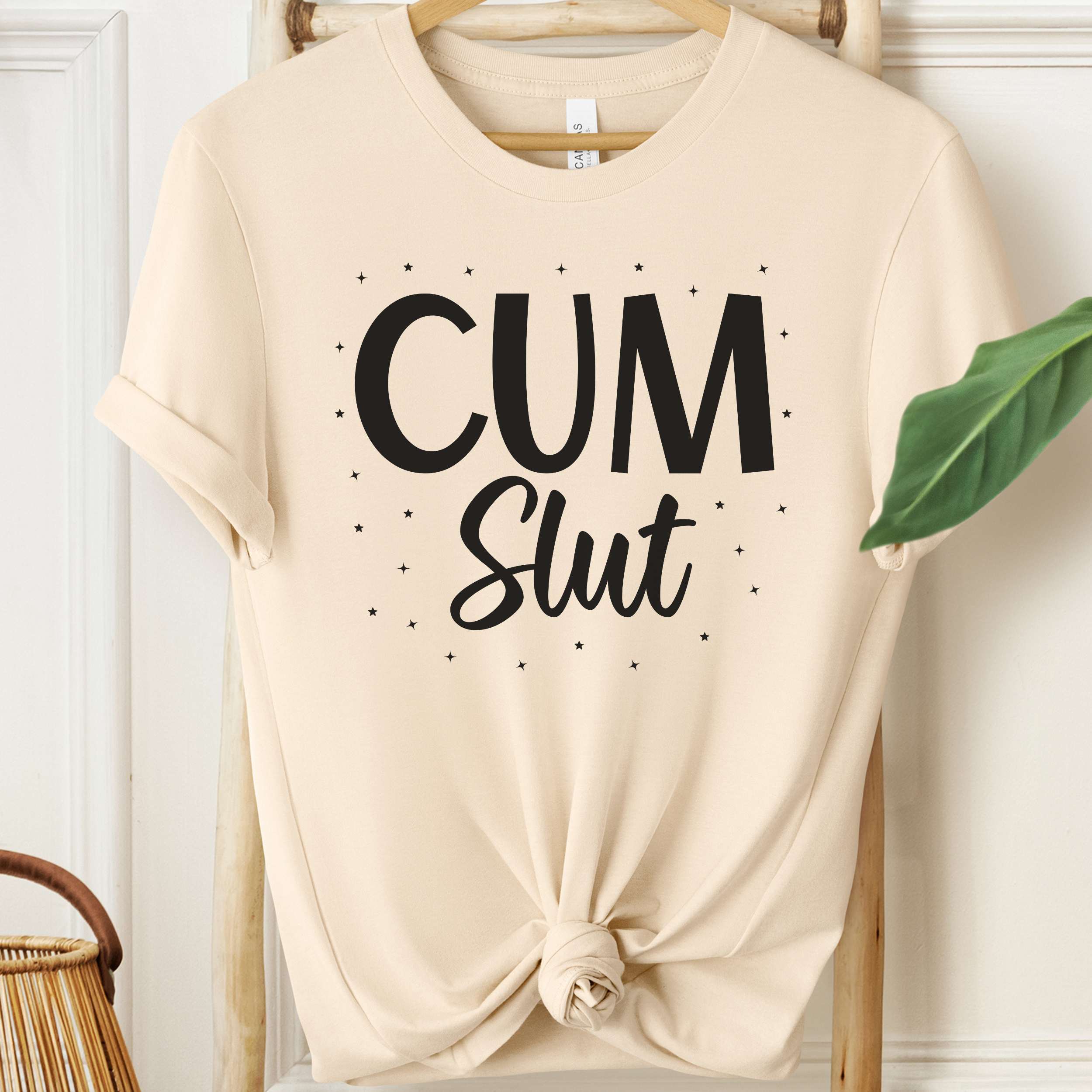 Beige t-shirt with 'Cum Slut' text on a hanger against a white wall.