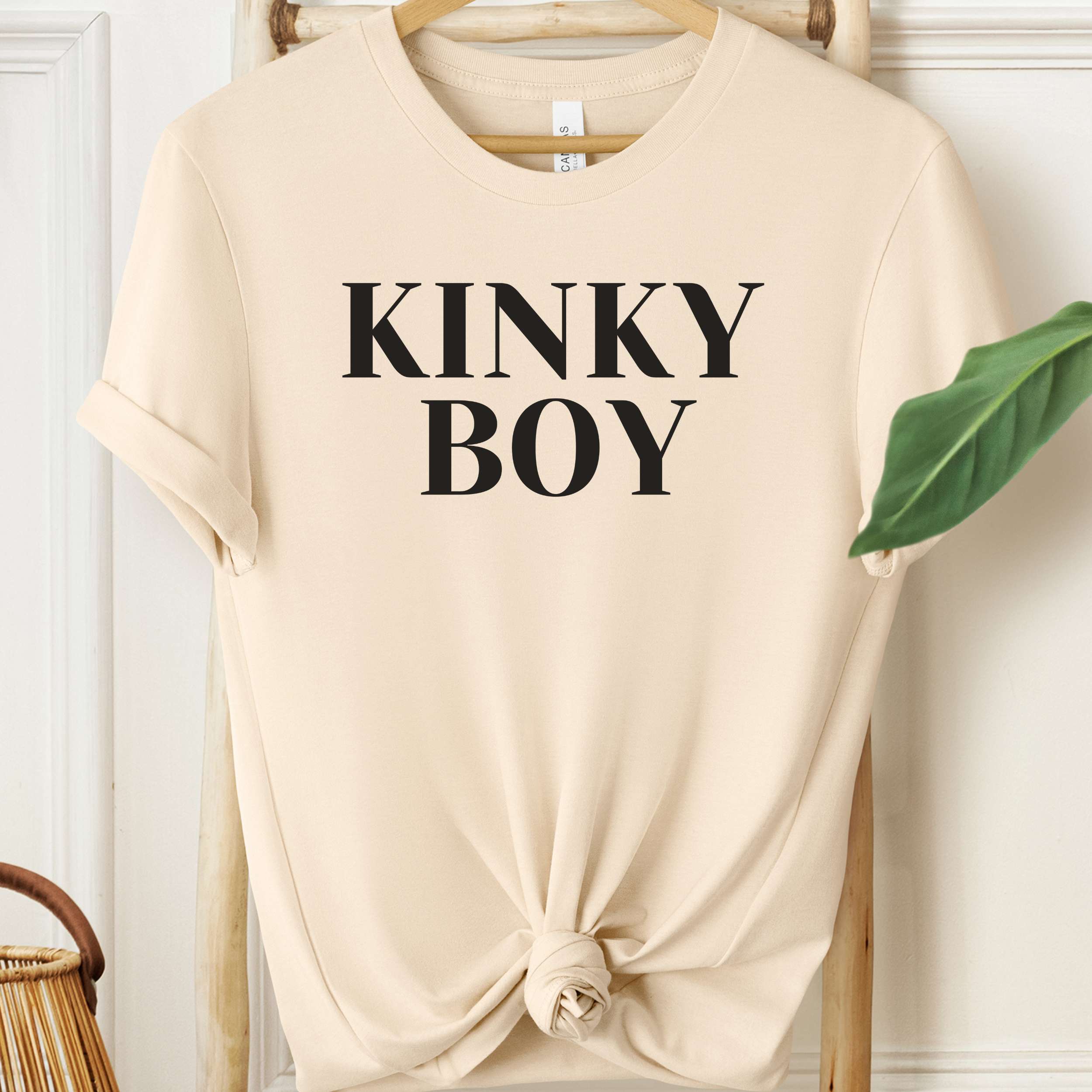 Beige t-shirt with 'KINKY BOY' text on a hanger against a white wall.