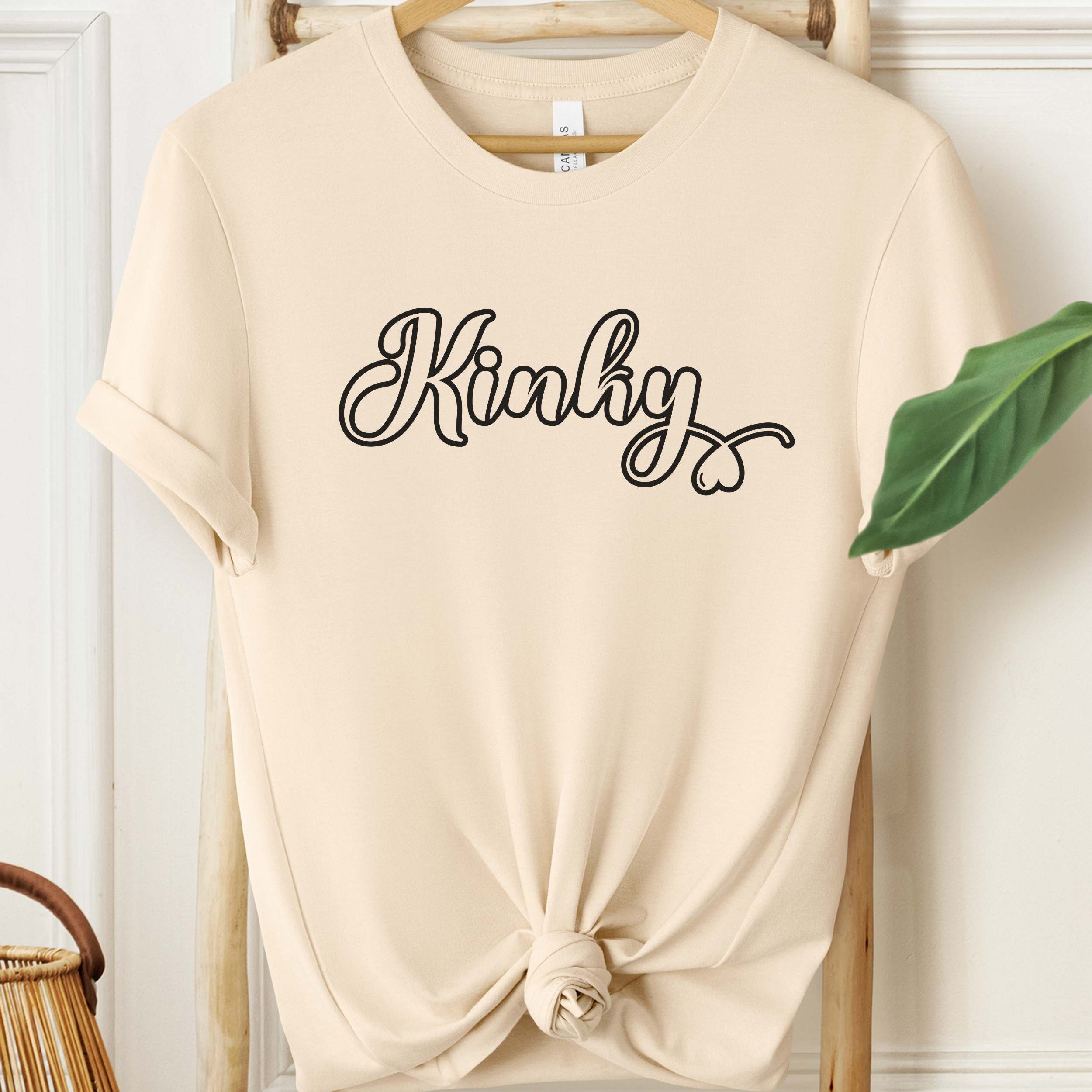 Beige t-shirt with 'Kinky' text on a hanger against a white wall.