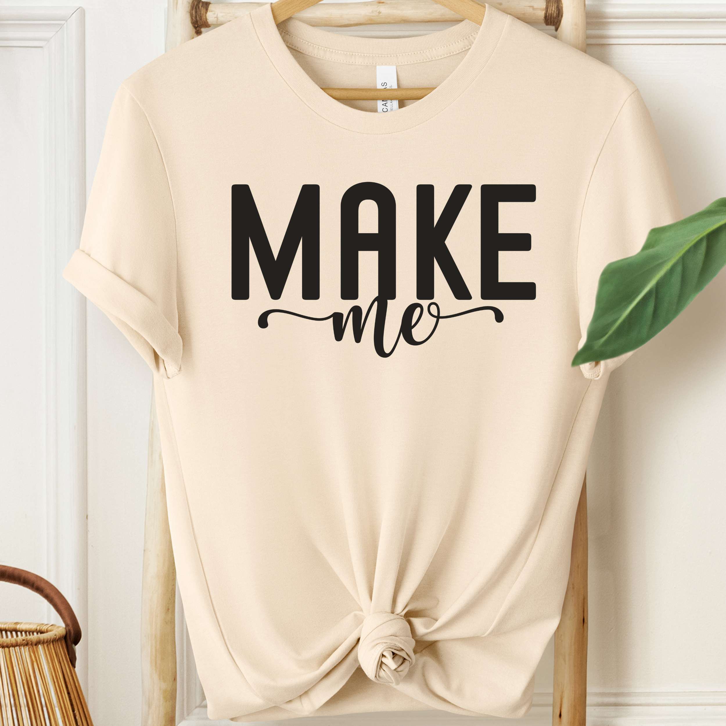 Beige t-shirt with 'MAKE me' text on a hanger against a white wall.