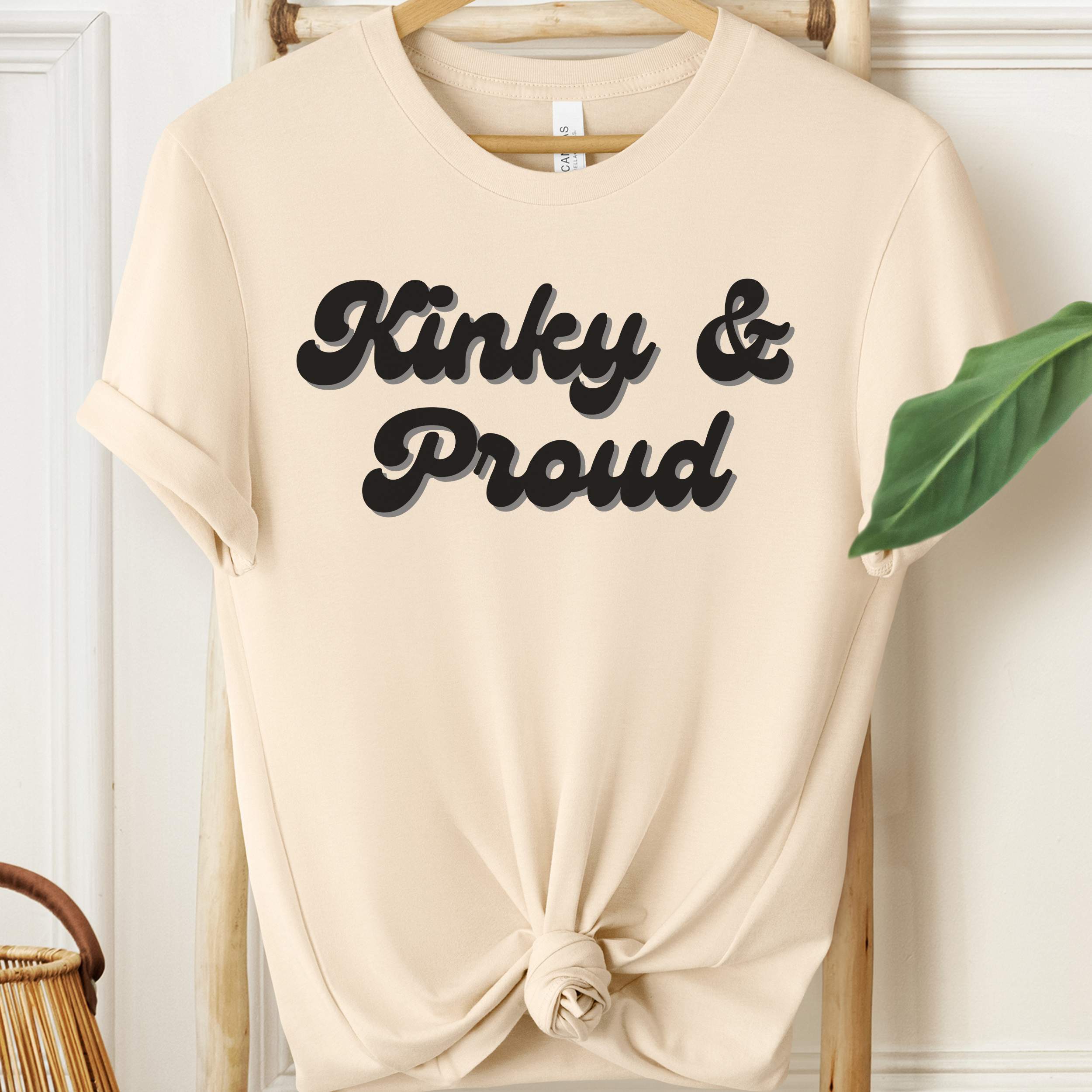 Beige t-shirt with 'Kinky & Proud' text on a hanger against a white wall.
