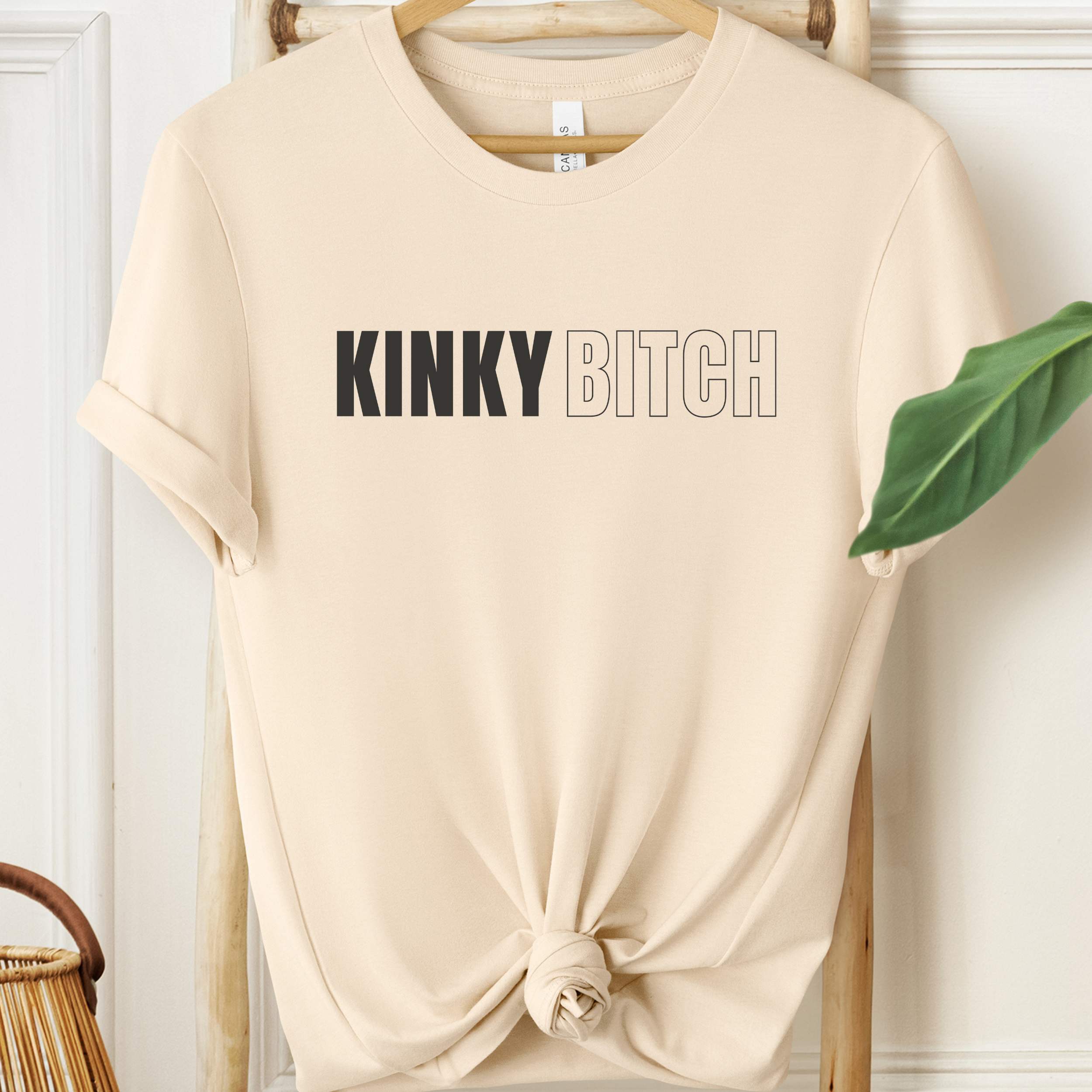 Beige t-shirt with 'KINKY BITCH' text on a hanger against a white wall.