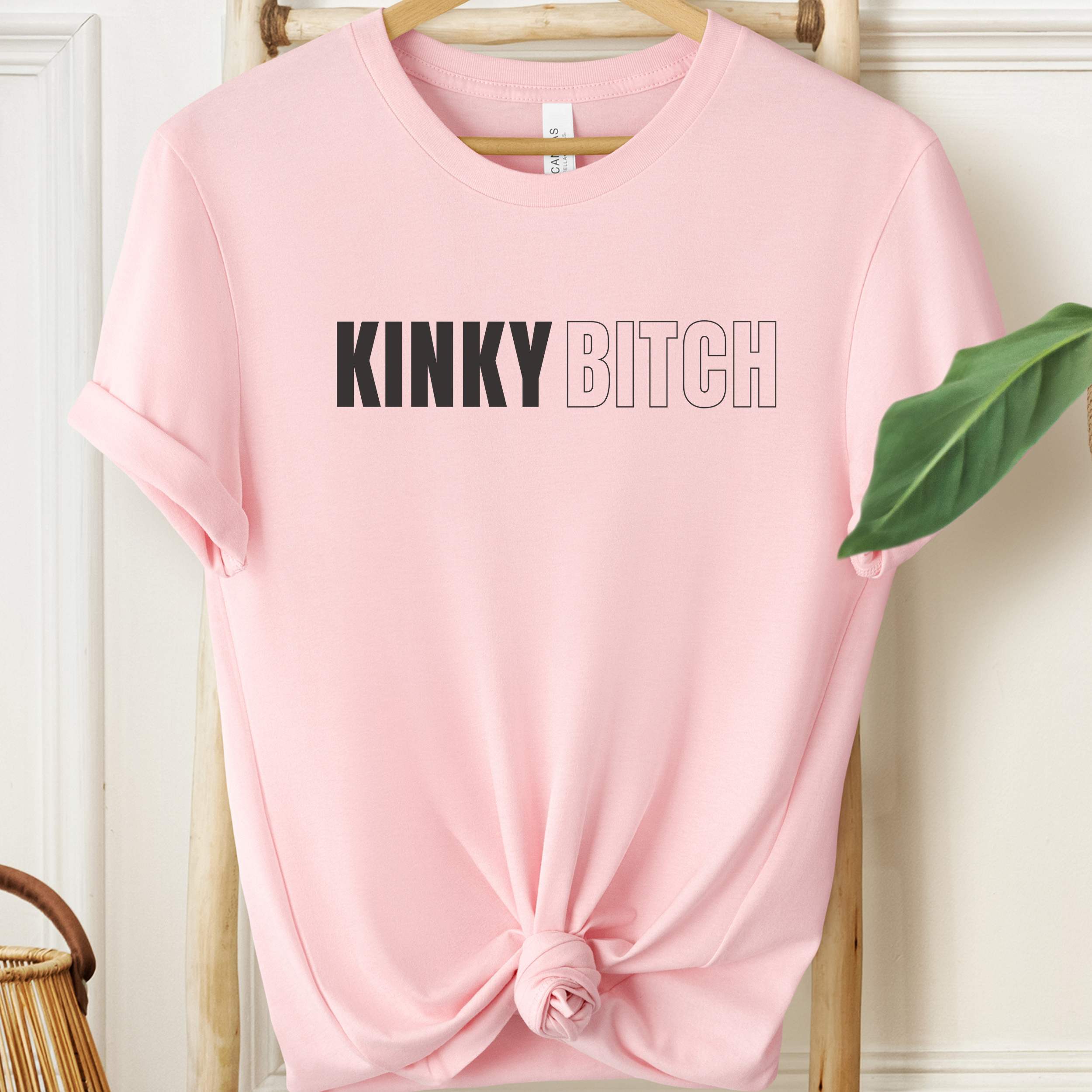 Pink t-shirt with 'KINKY BITCH' text on a hanger against a white wall.
