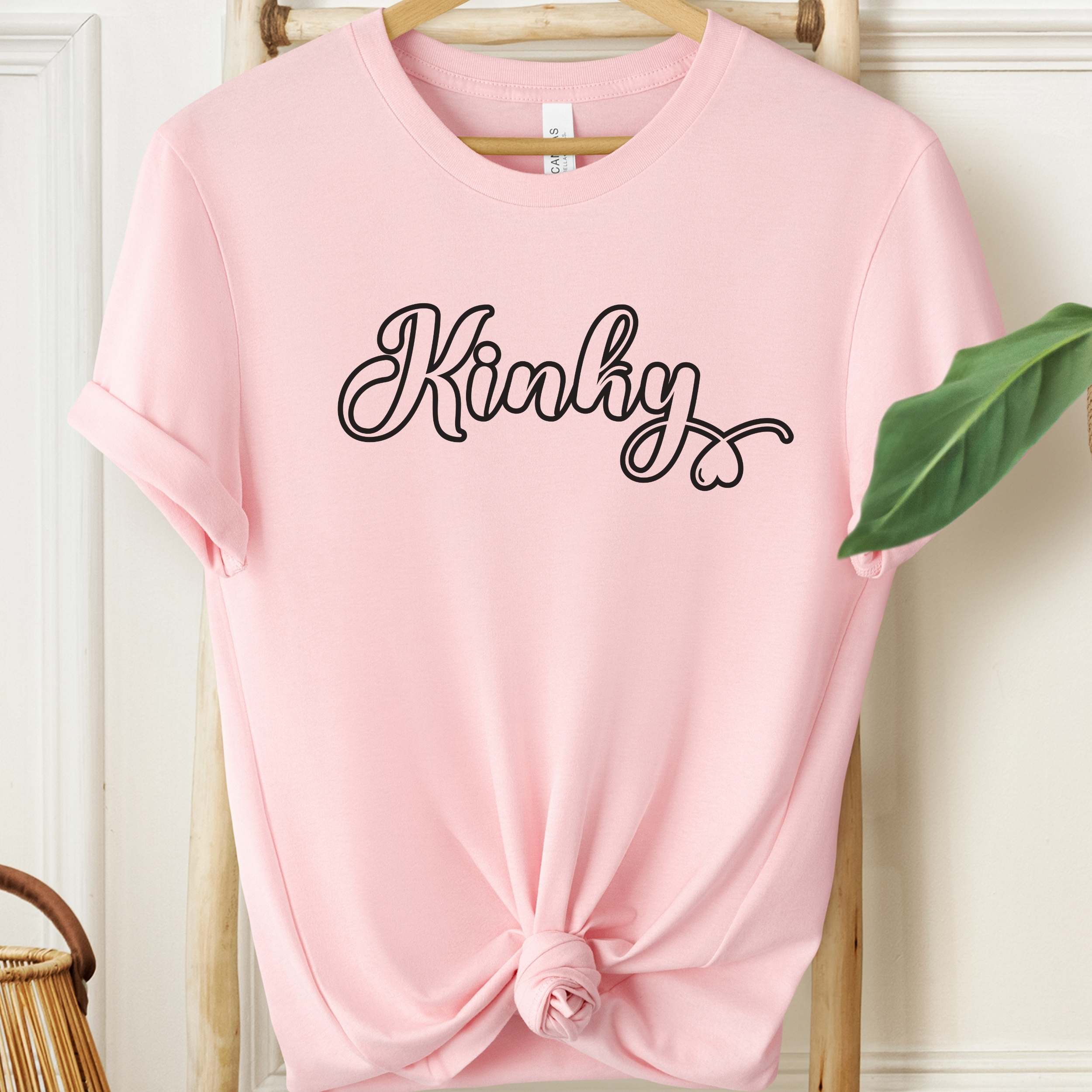 Pink t-shirt with 'Kinky' text on a hanger against a white wall.