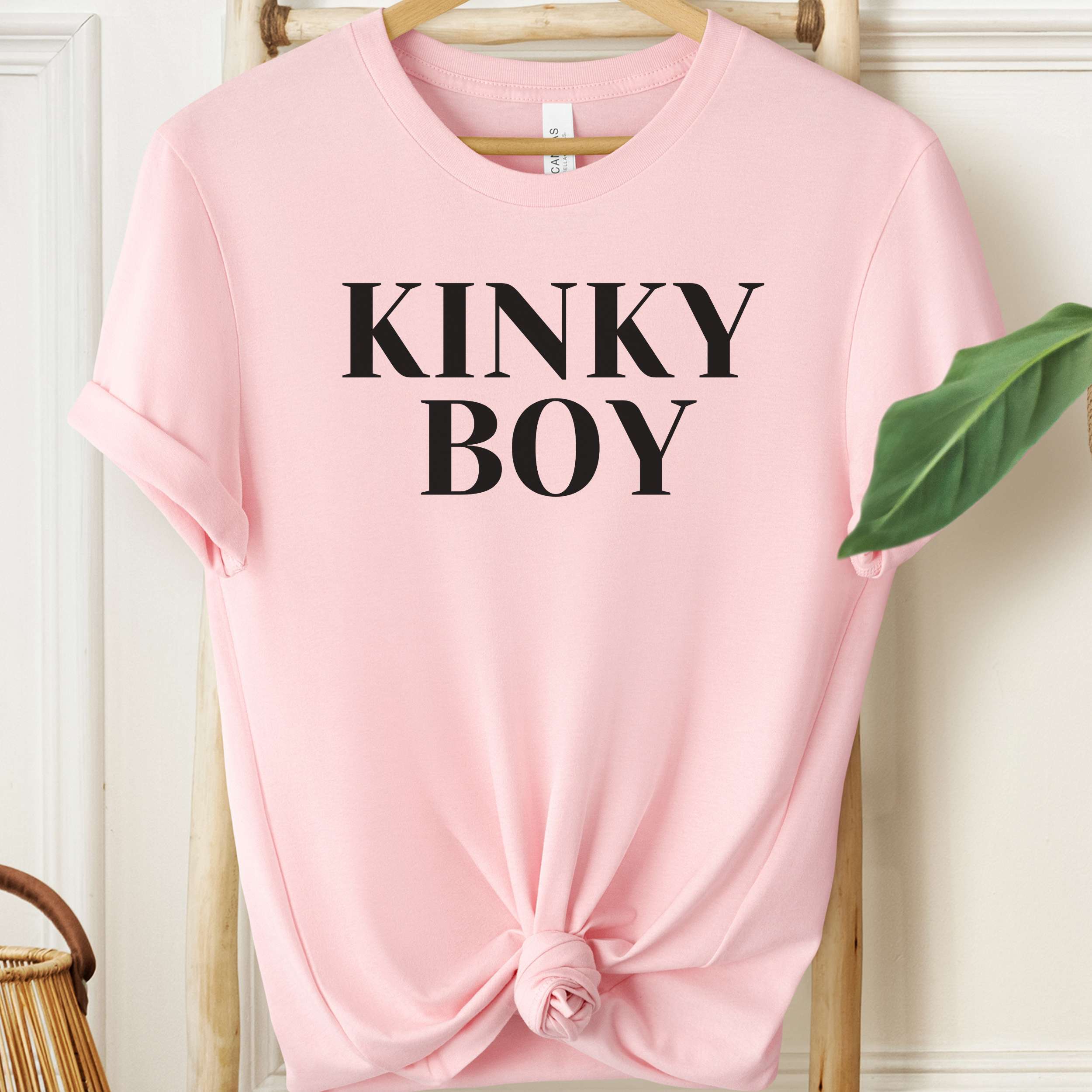 Pink t-shirt with 'KINKY BOY' text on a hanger against a white wall.