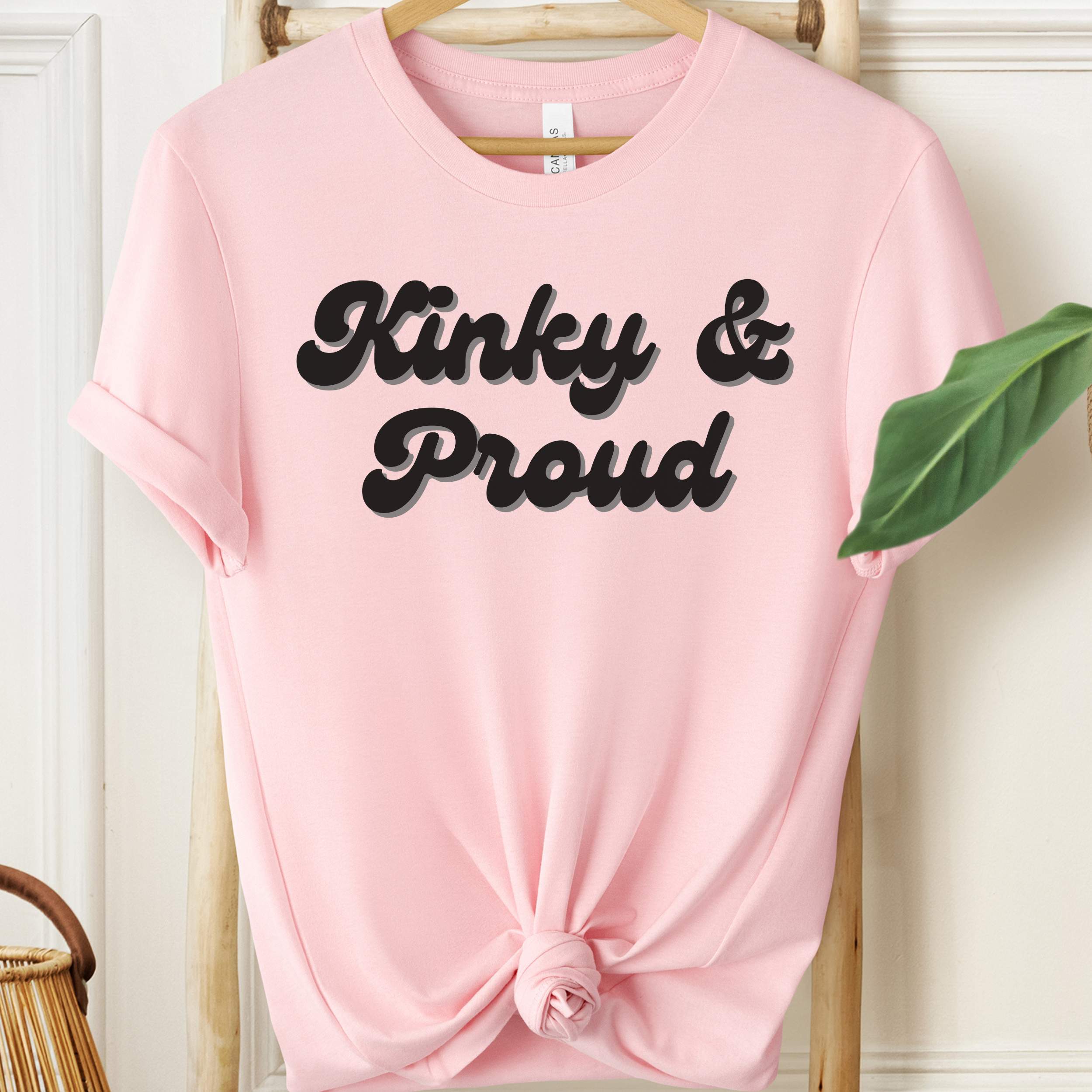 Pink t-shirt with 'Kinky & Proud' text on a hanger against a white wall.