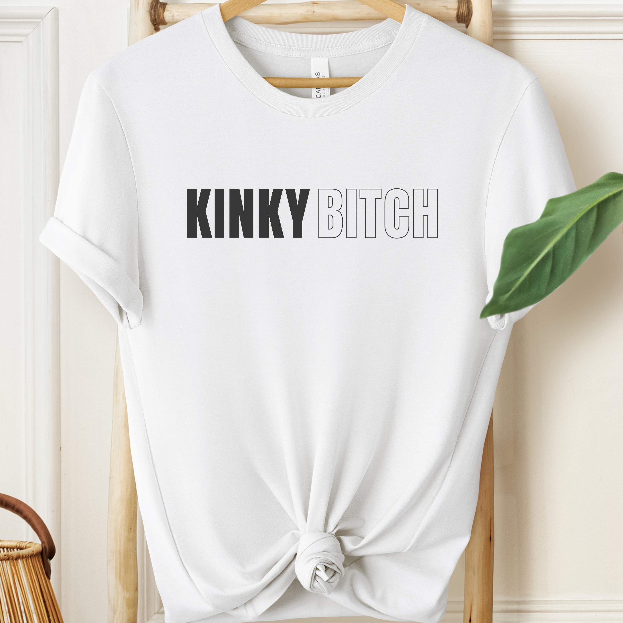 White t-shirt with 'KINKY BITCH' text on a hanger against a neutral background