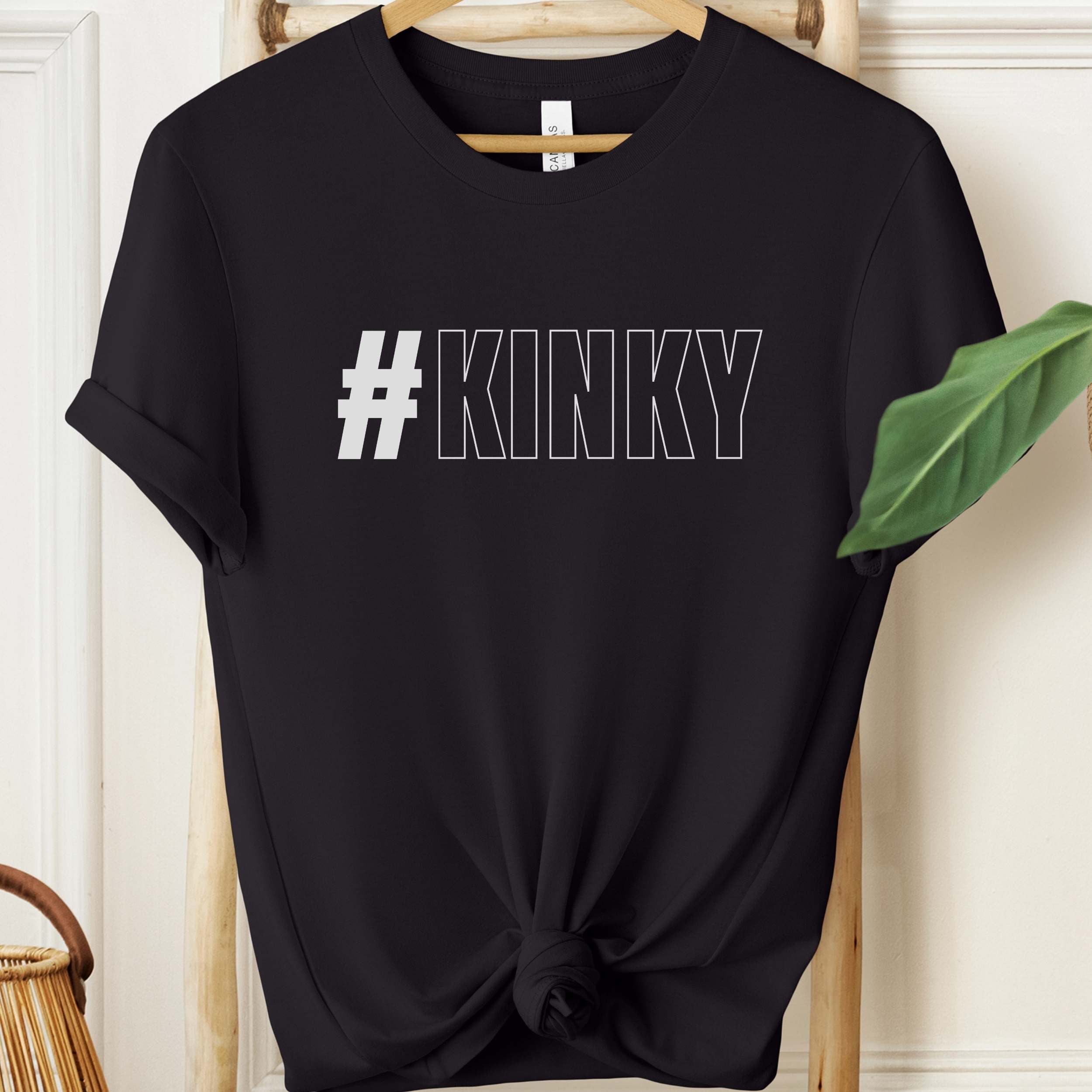 Black t-shirt with '#KINKY' text on a hanger against a white wall.