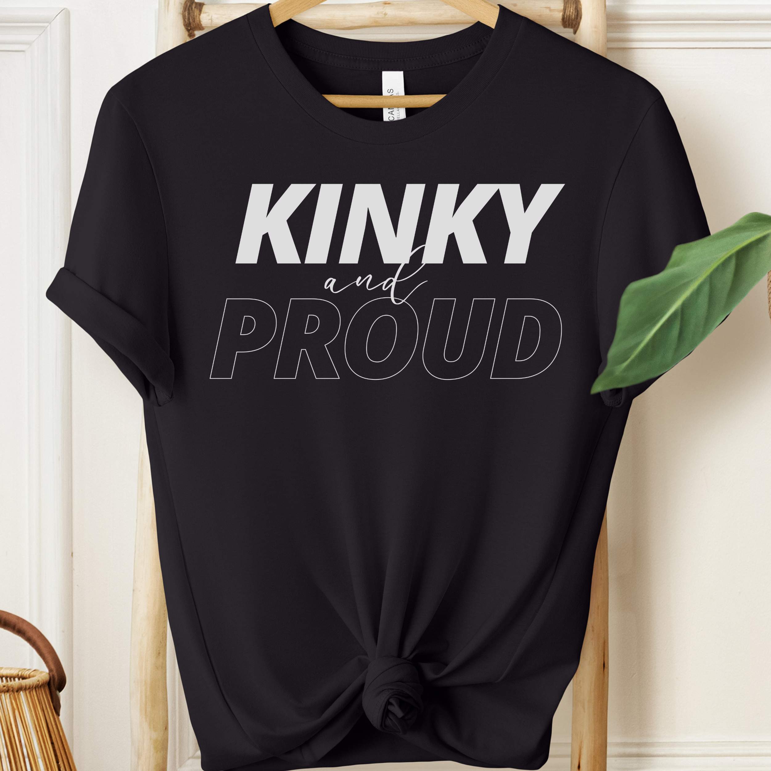 Black t-shirt with 'Kinky and Proud' text on a hanger against a white wall.