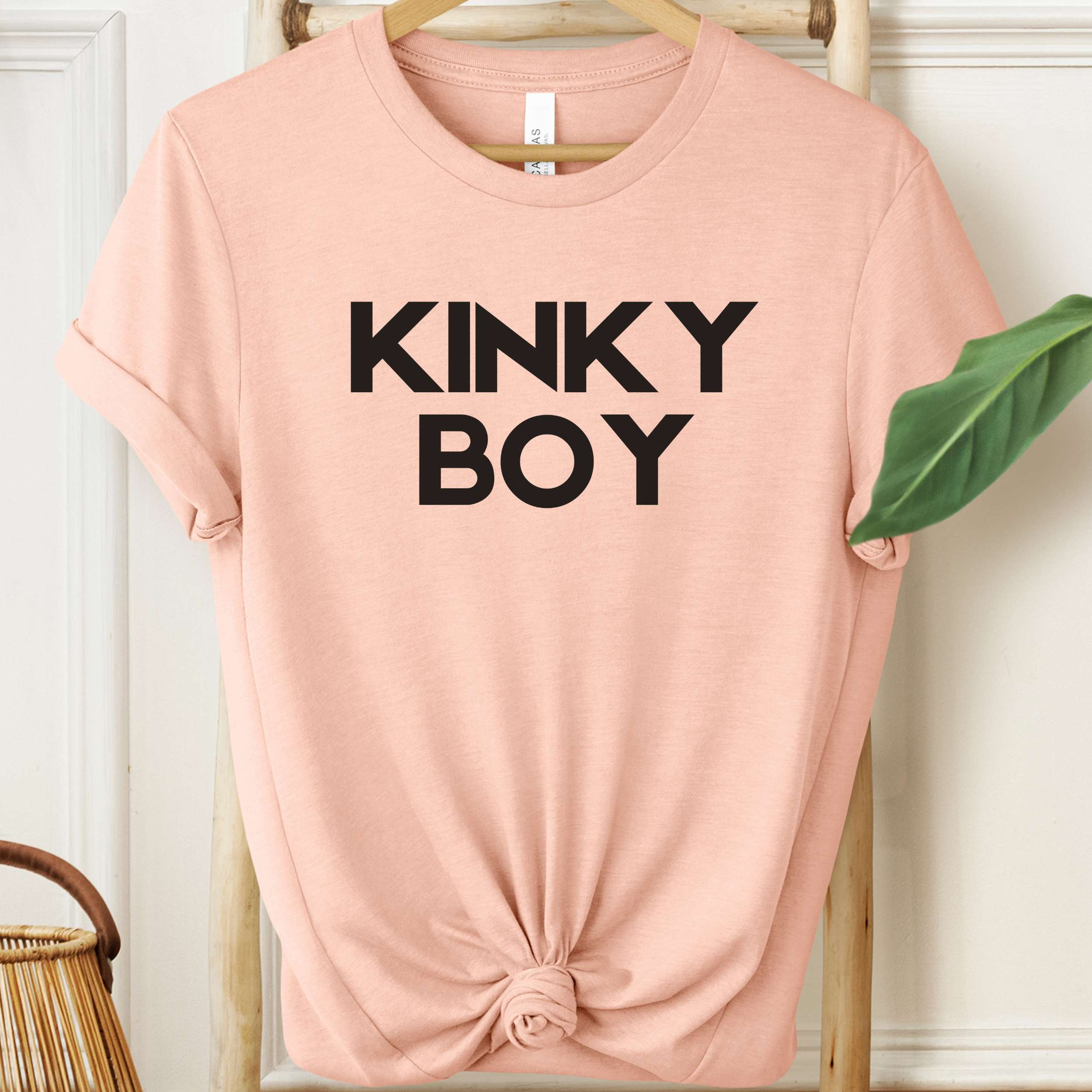 Pink t-shirt with 'KINKY BOY' text on a hanger against a white wall.