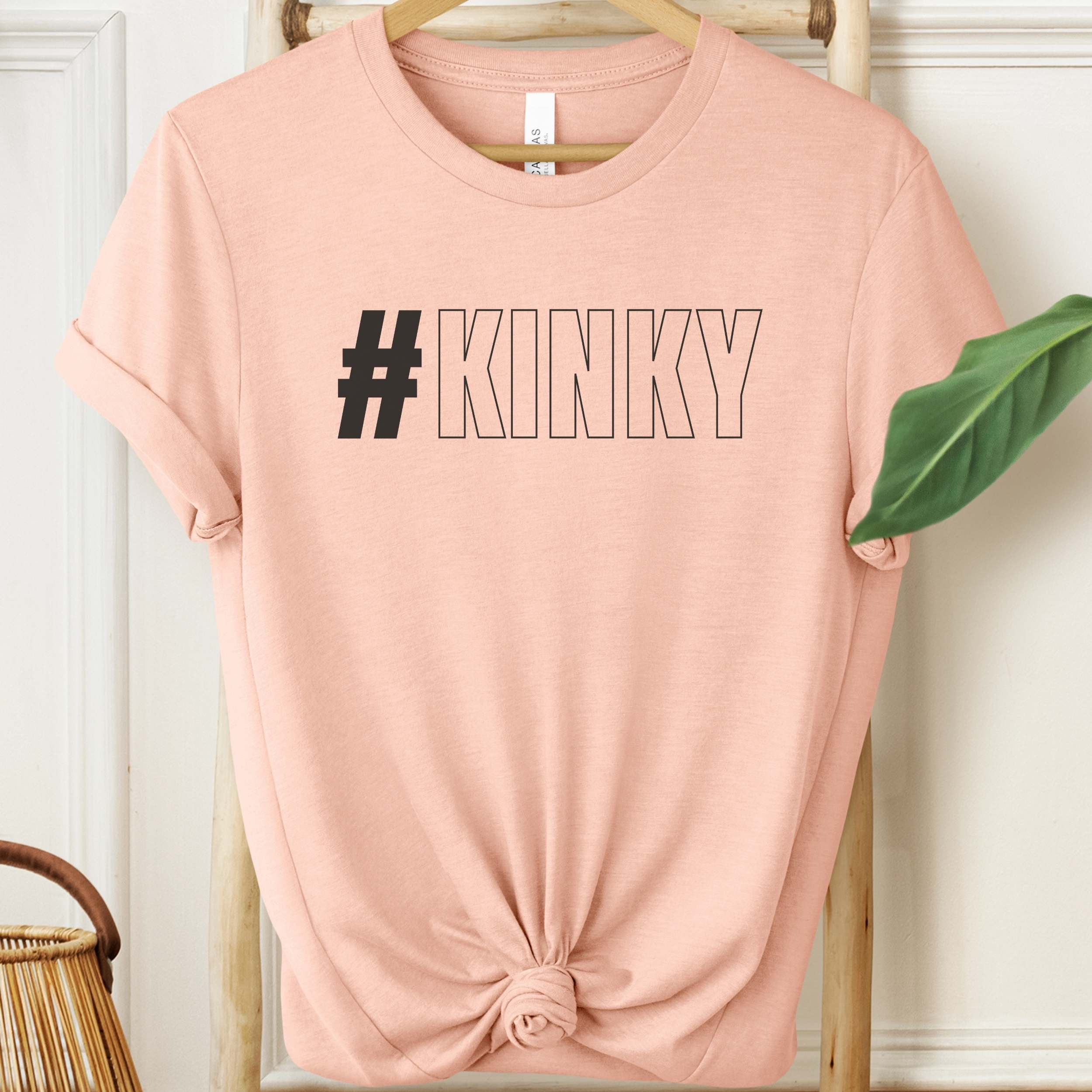 Pink t-shirt with '#KINKY' text on a hanger against a white wall.