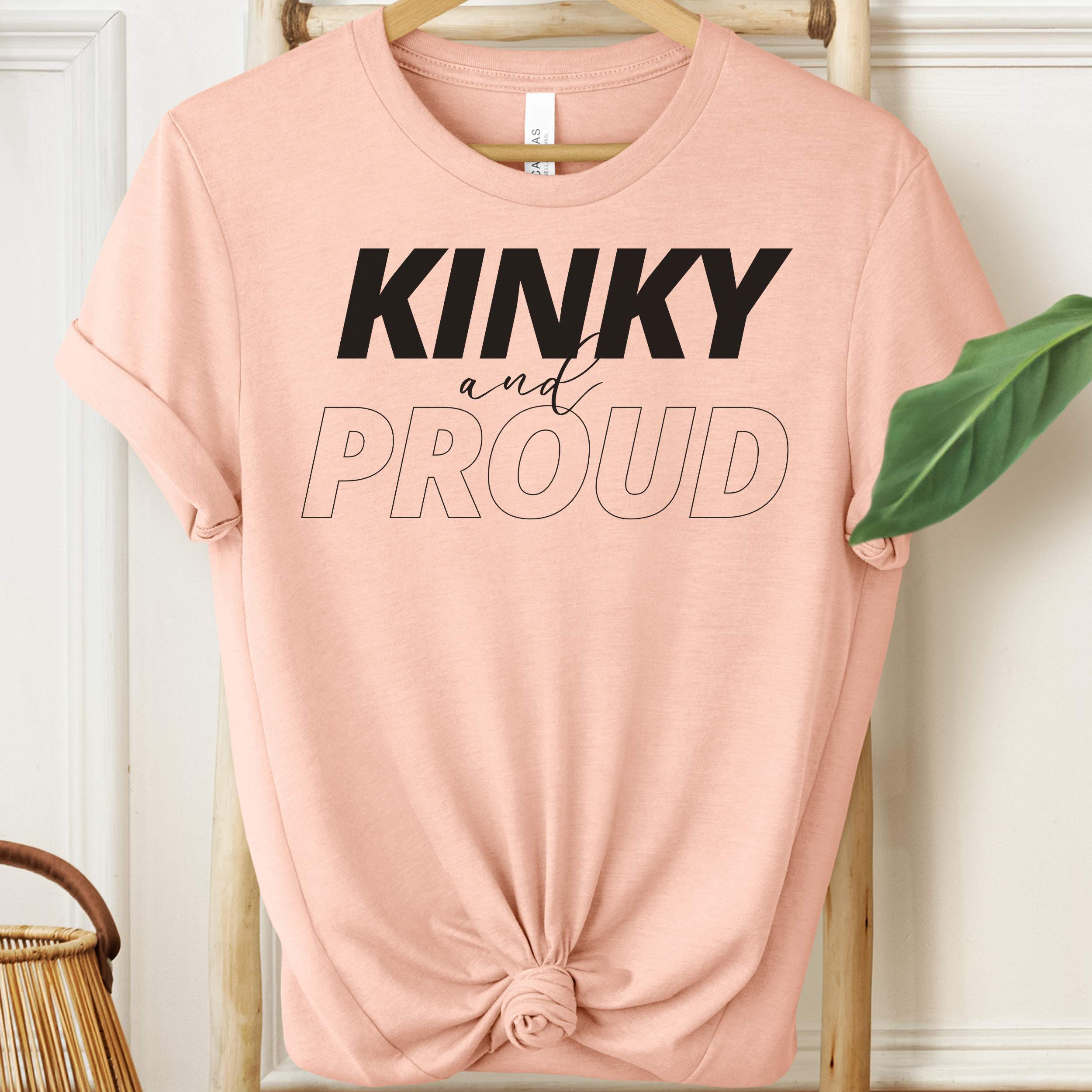 Pink t-shirt with 'KINKY and PROUD' text on a hanger against a white wall.