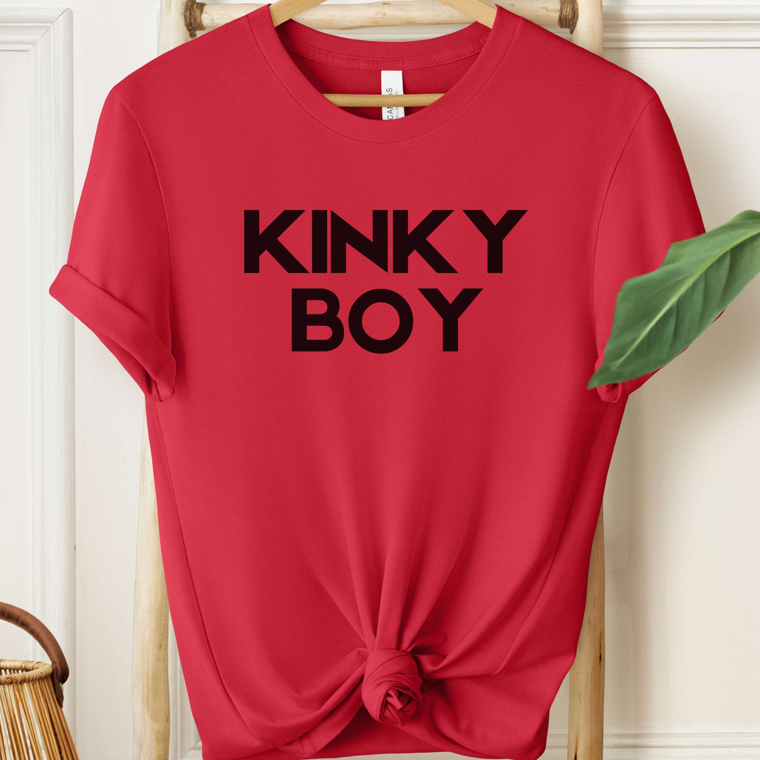 Red t-shirt with 'KINKY BOY' text on a hanger against a white wall.