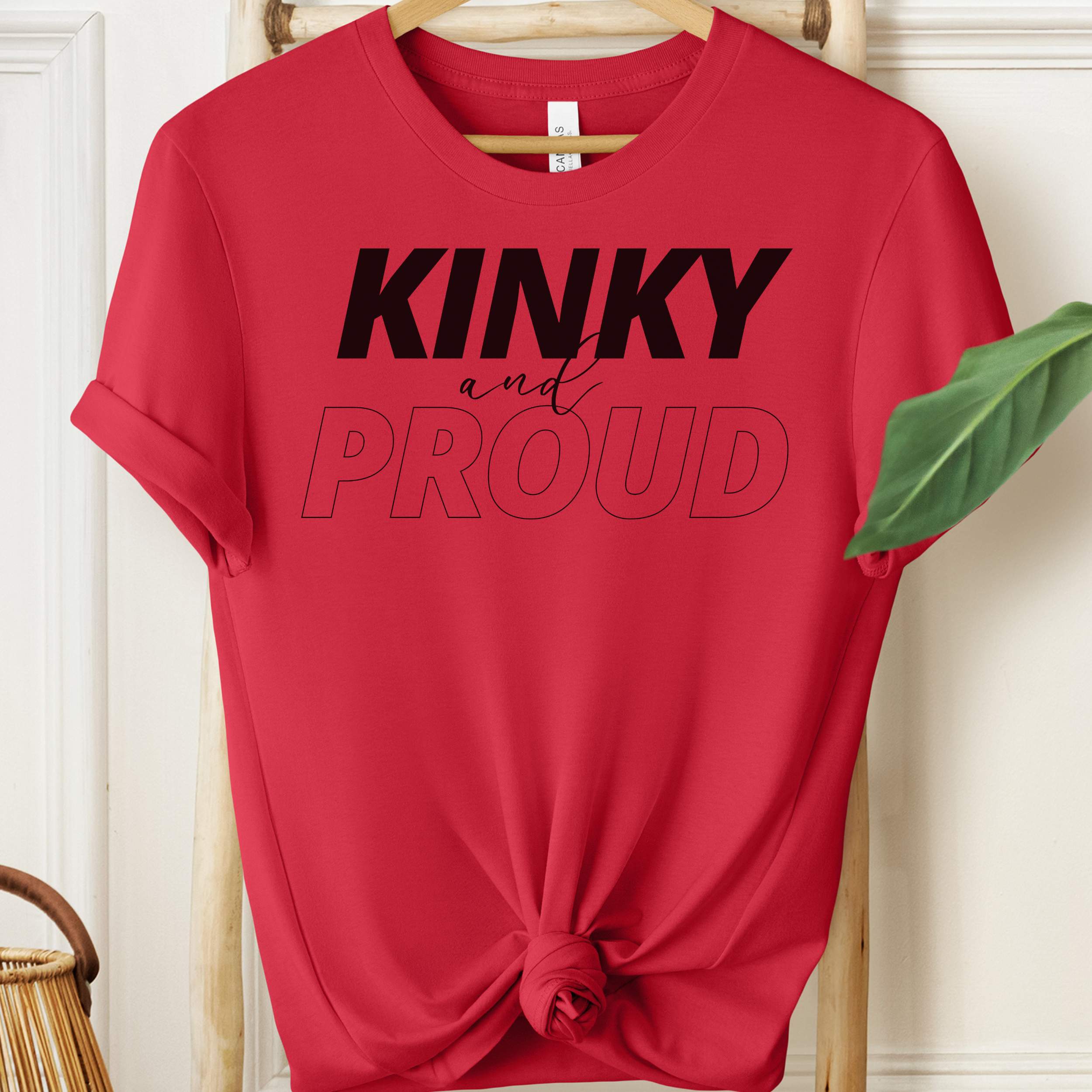 Red t-shirt with 'Kinky and Proud' text on a hanger against a white wall.